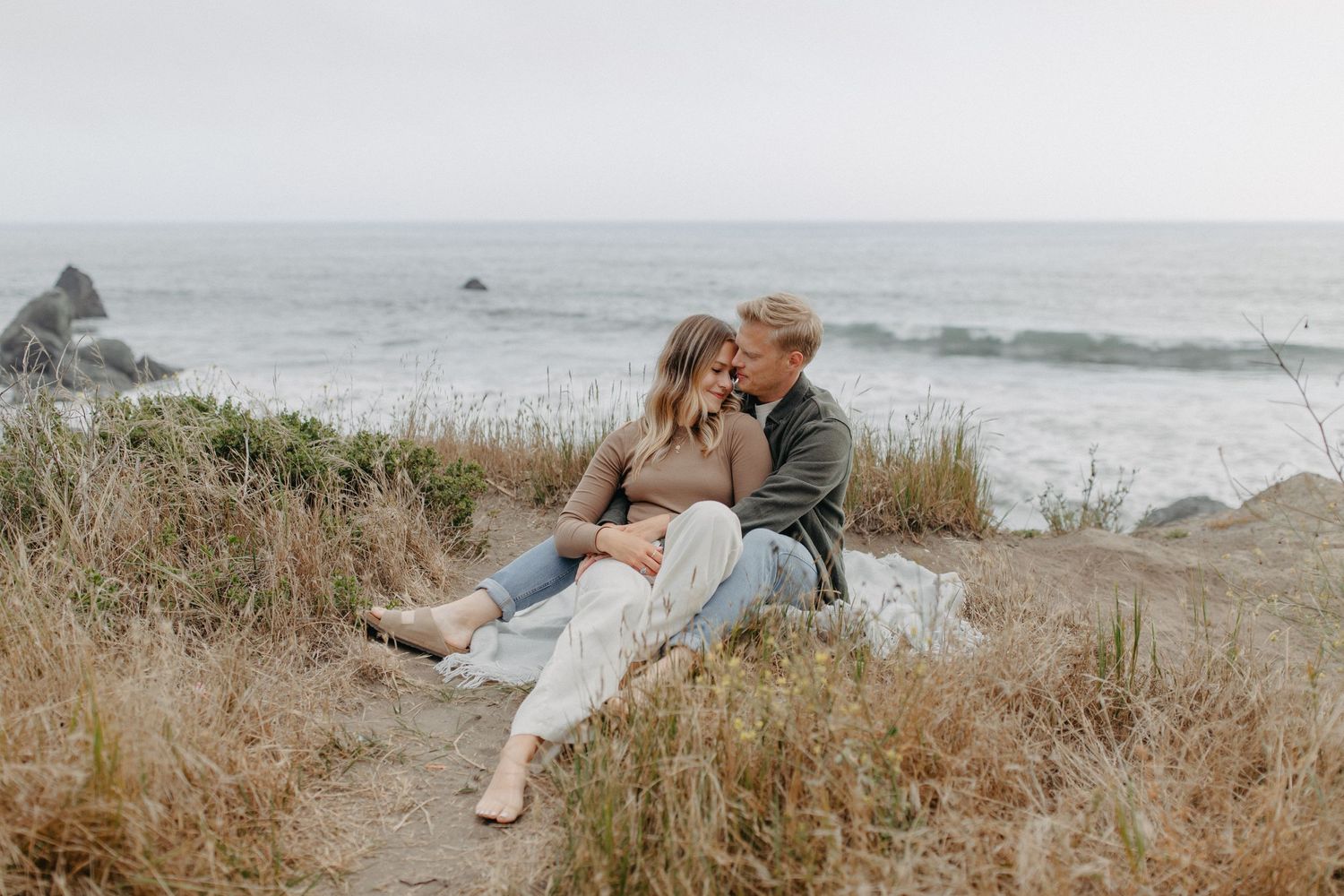 Romantic couple cuddling on scenic coastal cliffside with ocean waves and rocks in background.
