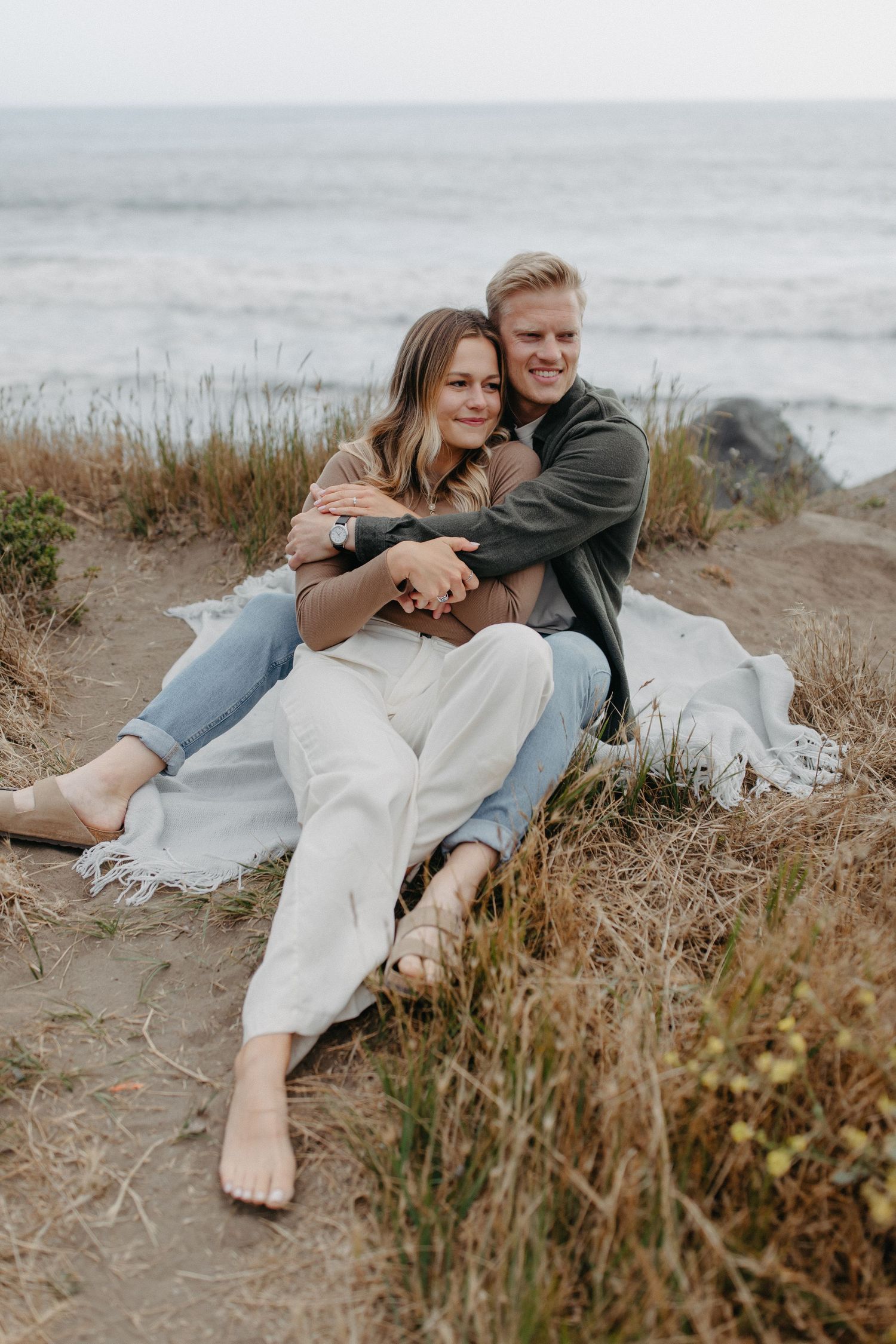 Couple in casual wear sharing tender moment while sitting on coastal path with ocean view.