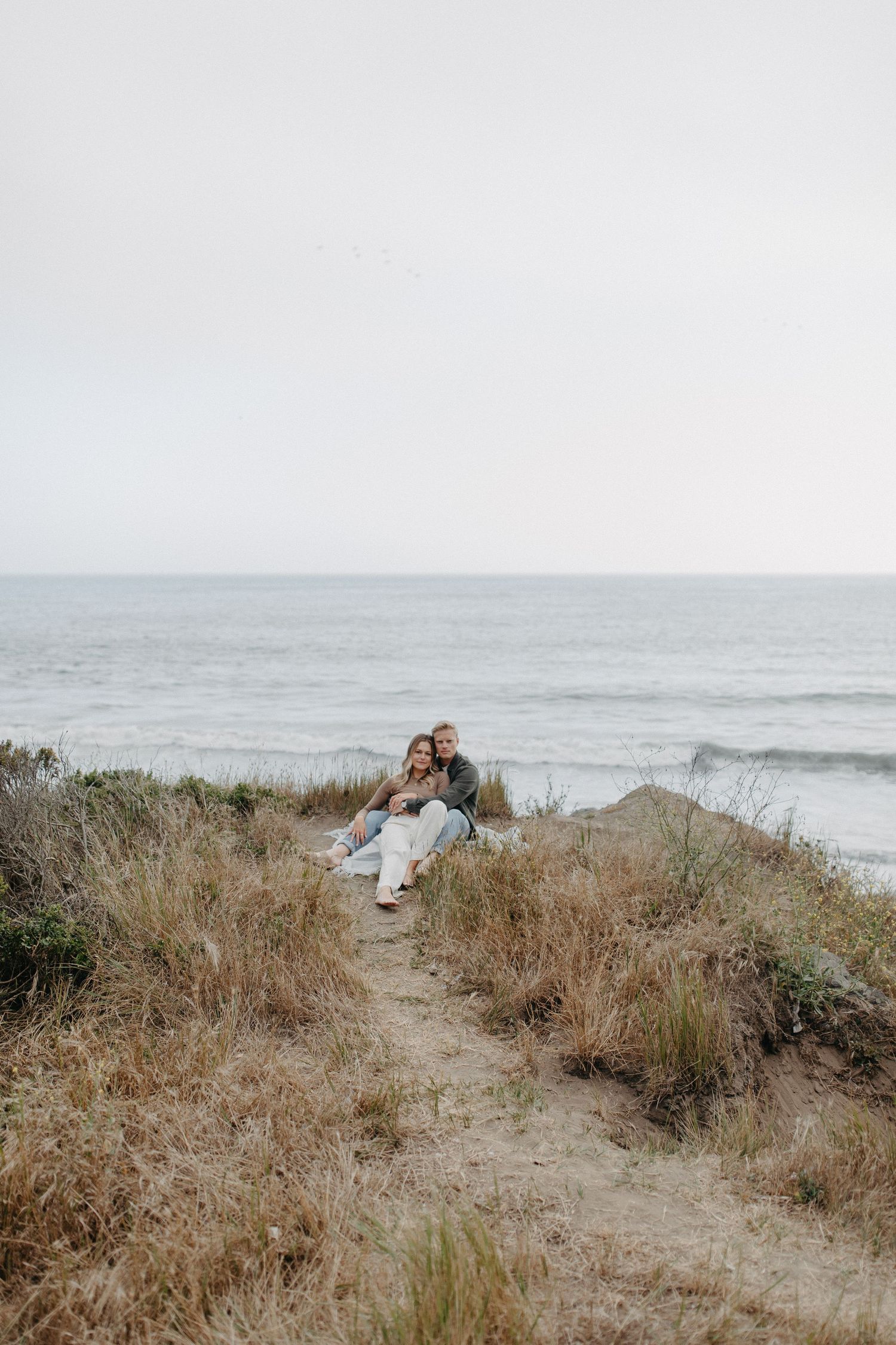 Distant view of couple sitting on coastal trail surrounded by dry grass with ocean backdrop.