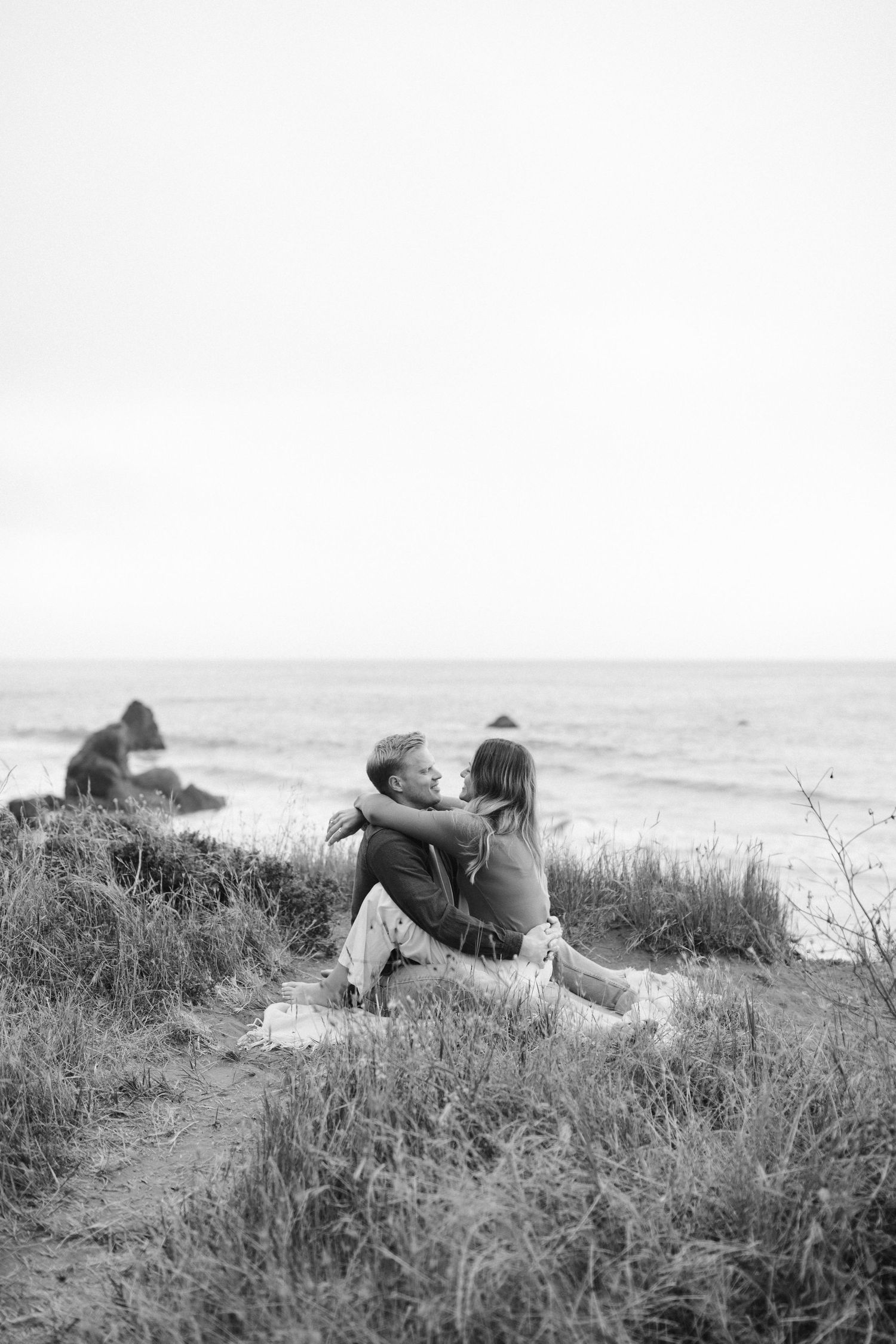 Black and white image of couple embracing on grassy oceanside cliff.