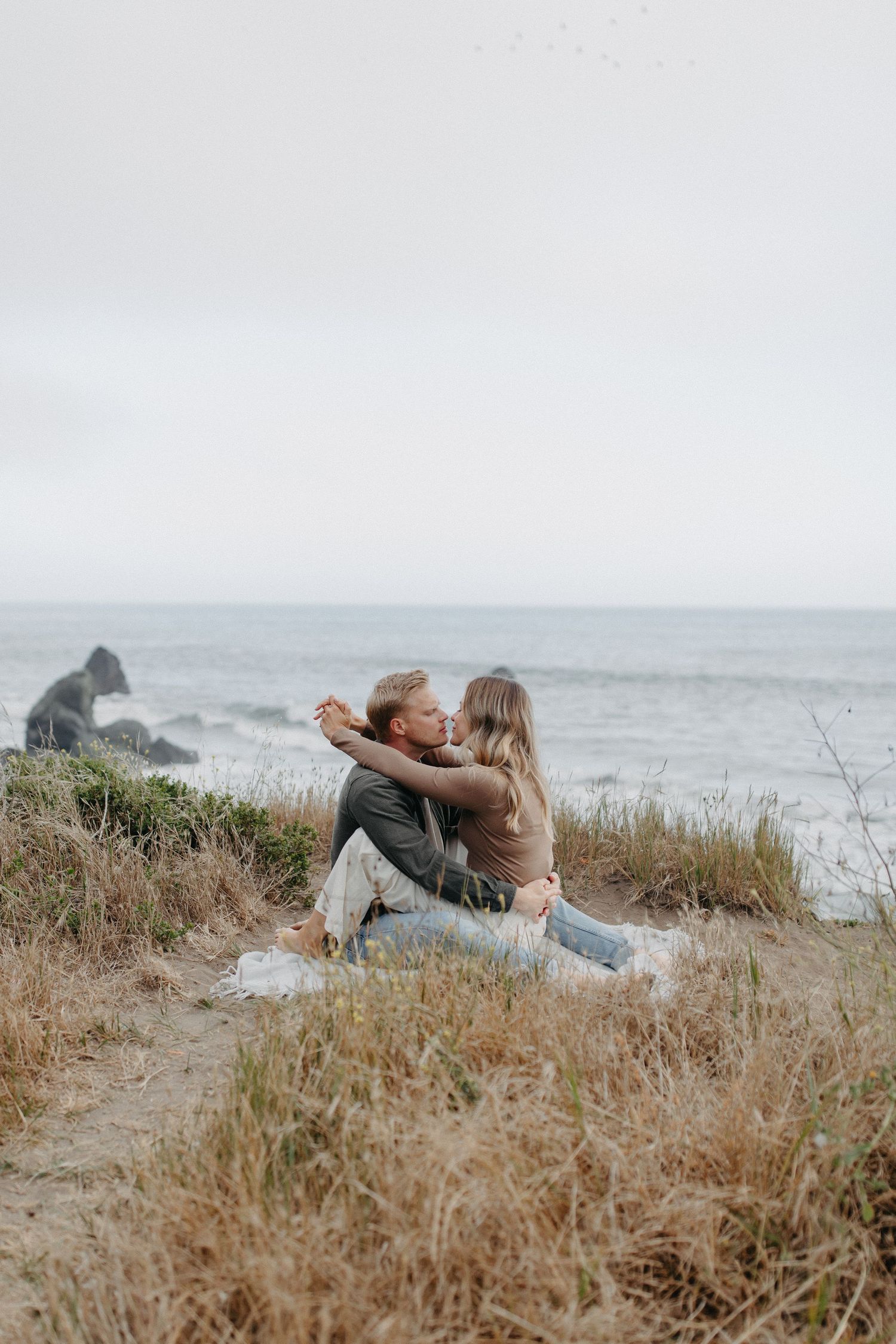 Intimate moment between couple on seaside cliff with ocean waves in background.