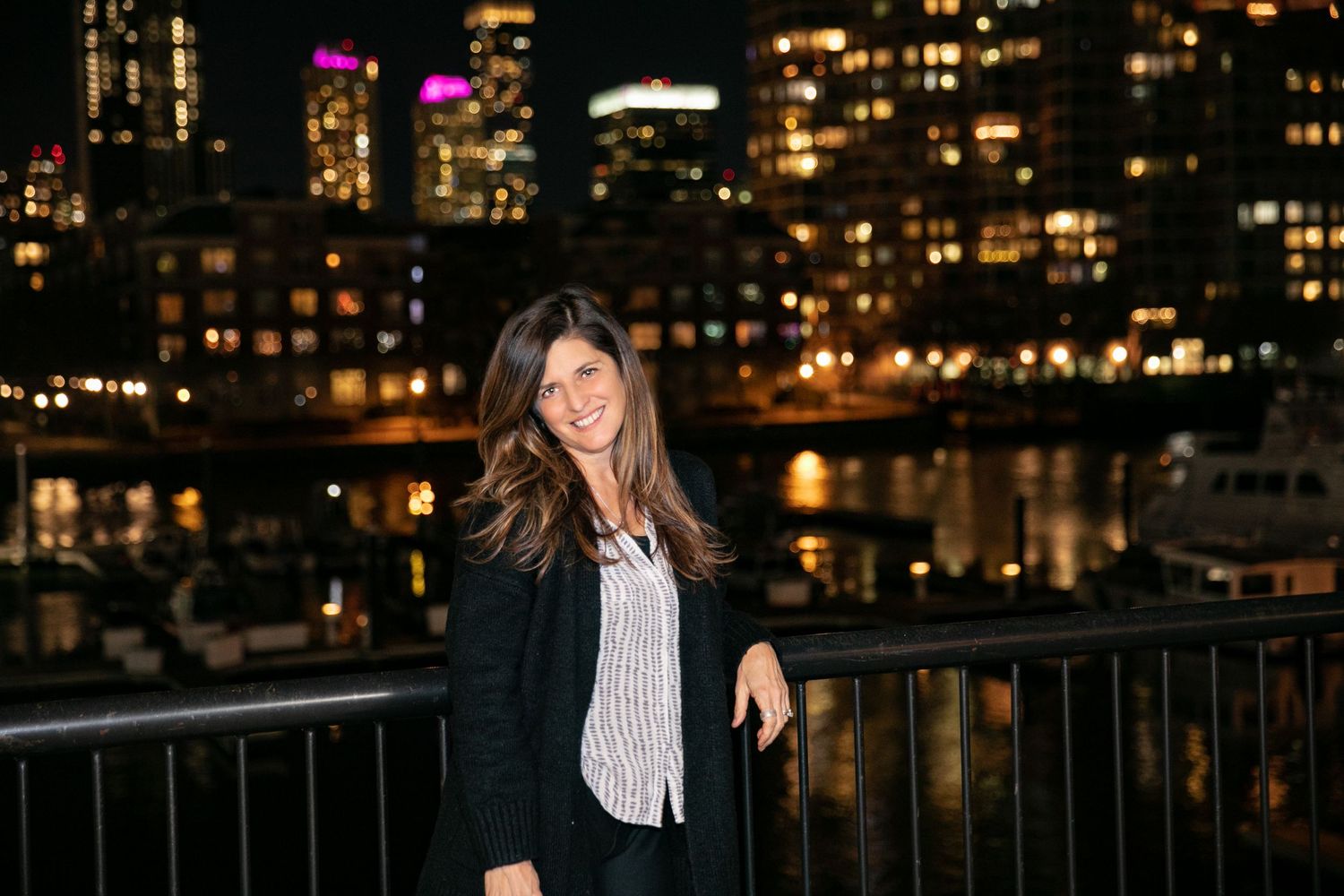 Kathy Emons in front of a cityscape at night time wearing a black cardigan, white patterned shirt, and black slacks.