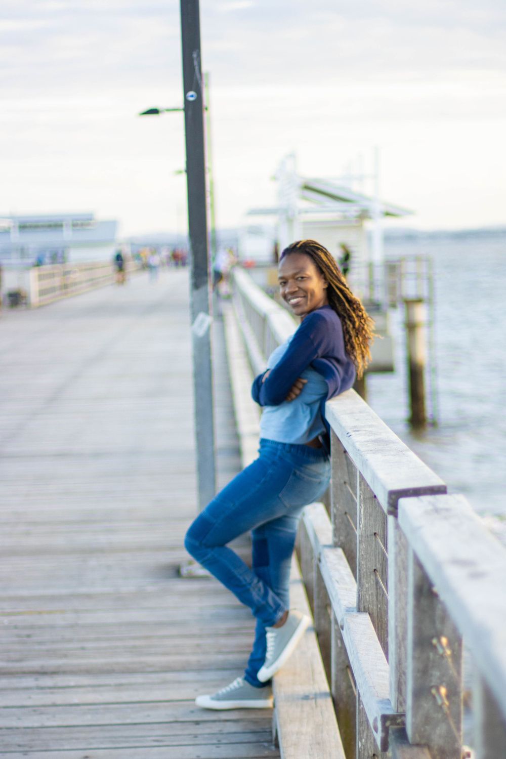 A casual portrait of someone in blue denim leaning against a pier railing with water in the background.