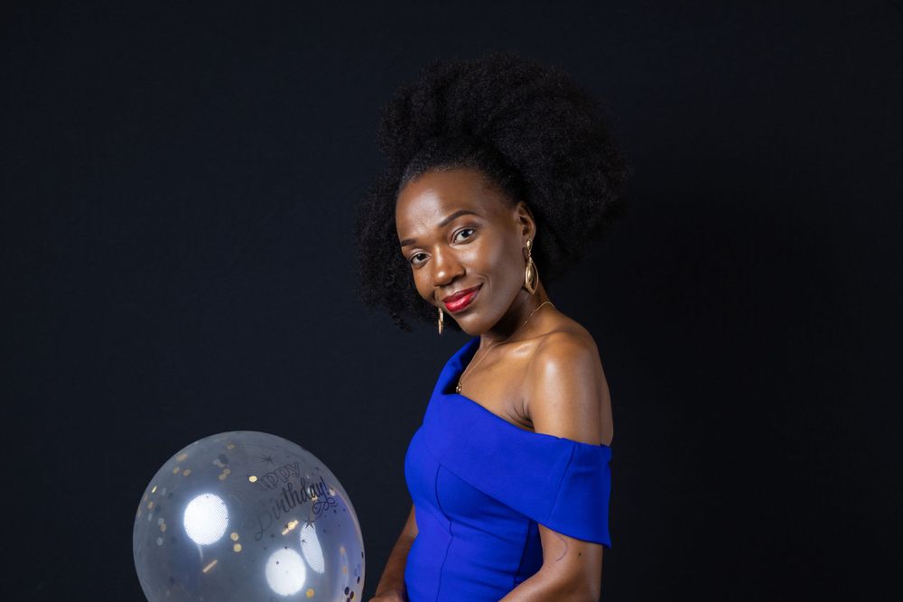 A person in a royal blue off-shoulder dress poses against a dark background with a clear balloon.