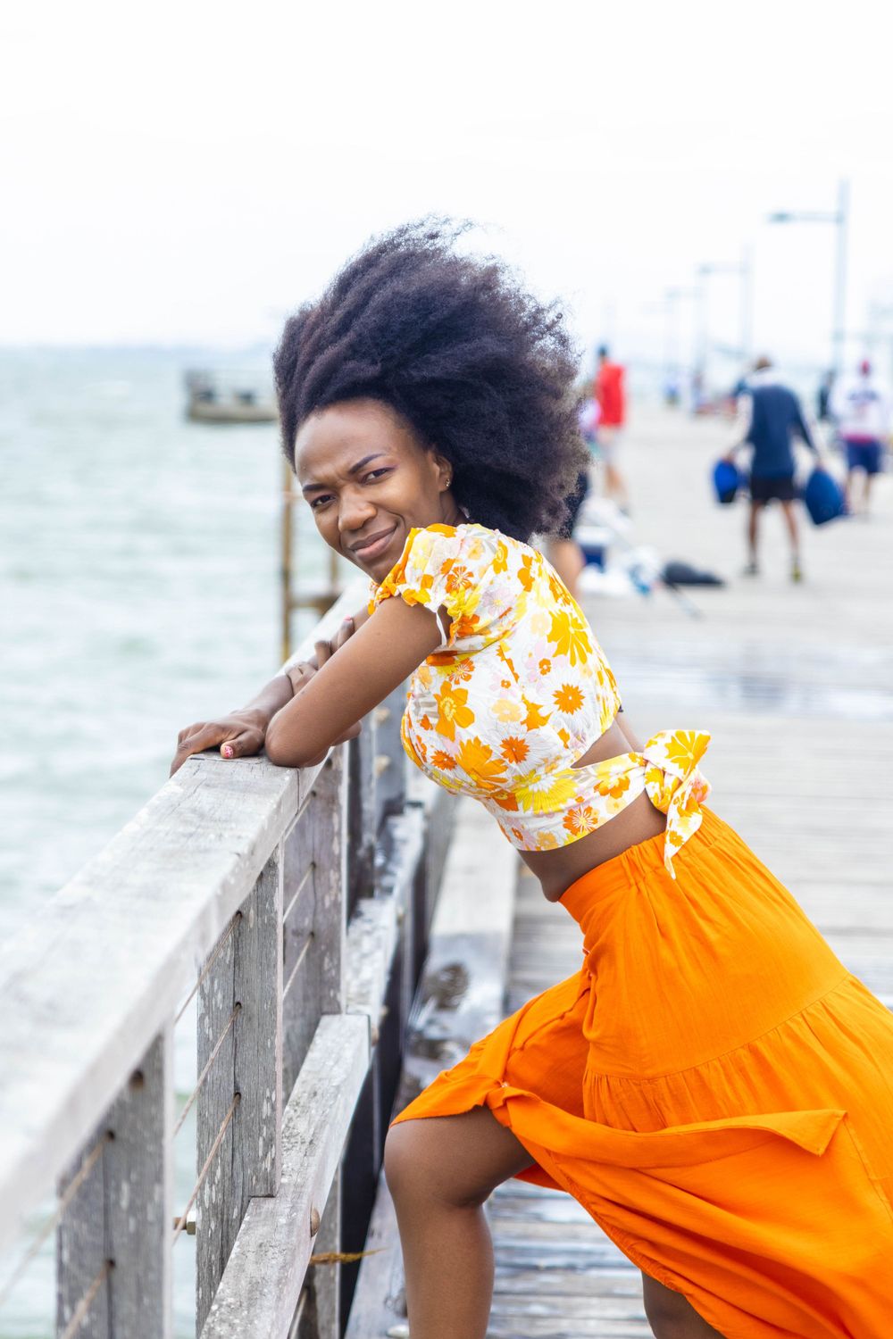 Person in floral top and orange skirt posing playfully on oceanfront pier.