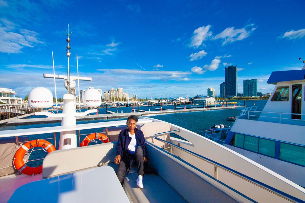 A person relaxes on the deck of a boat with a cityscape visible across the harbor.