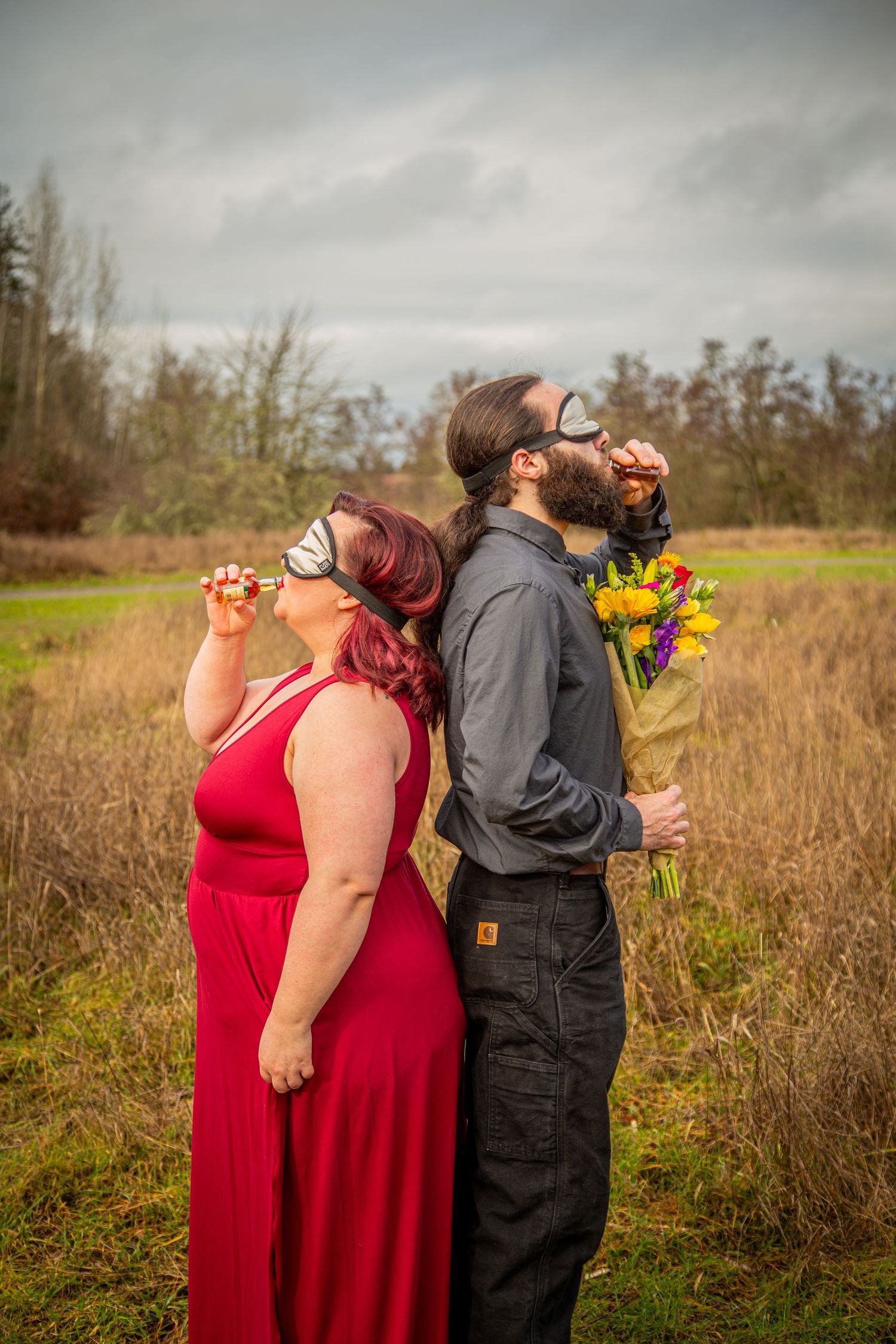 Two people stand back-to-back outdoors, one in red dress and one in dark clothing, both looking upward with flowers.