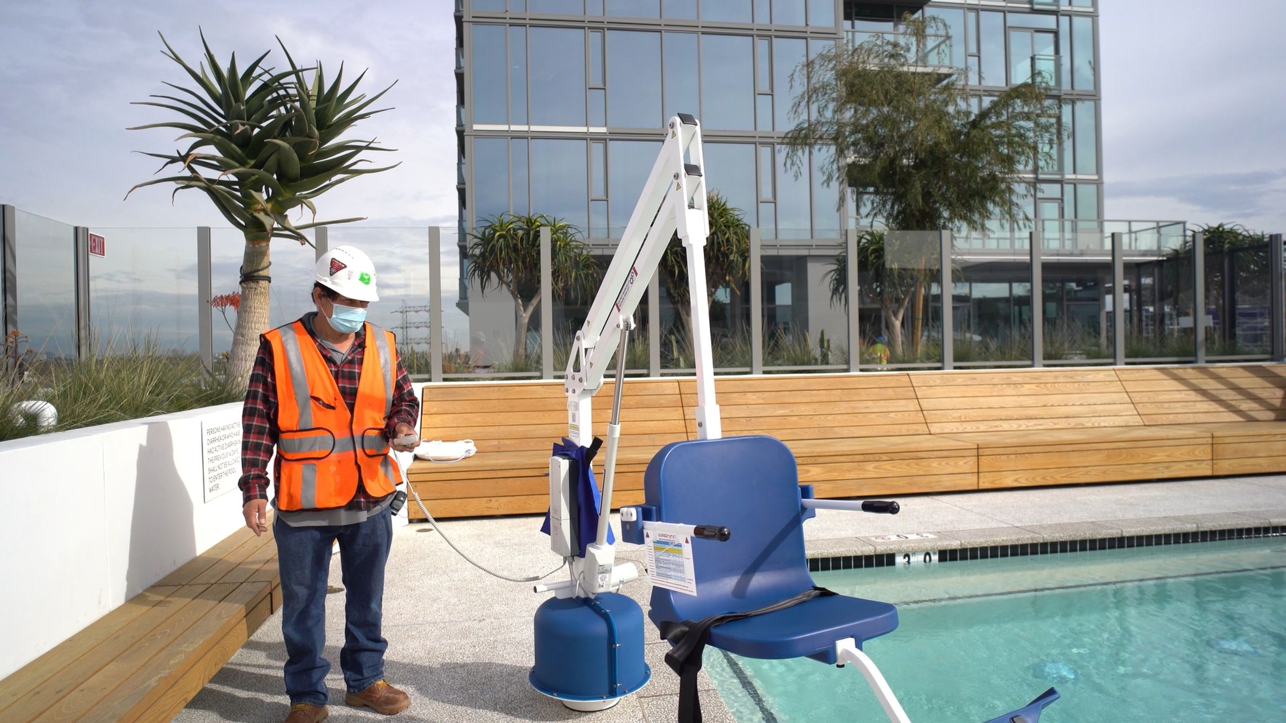 Safety inspector wearing a hard hat and high-visibility vest testing mechanical pool equipment at a rooftop swimming pool.