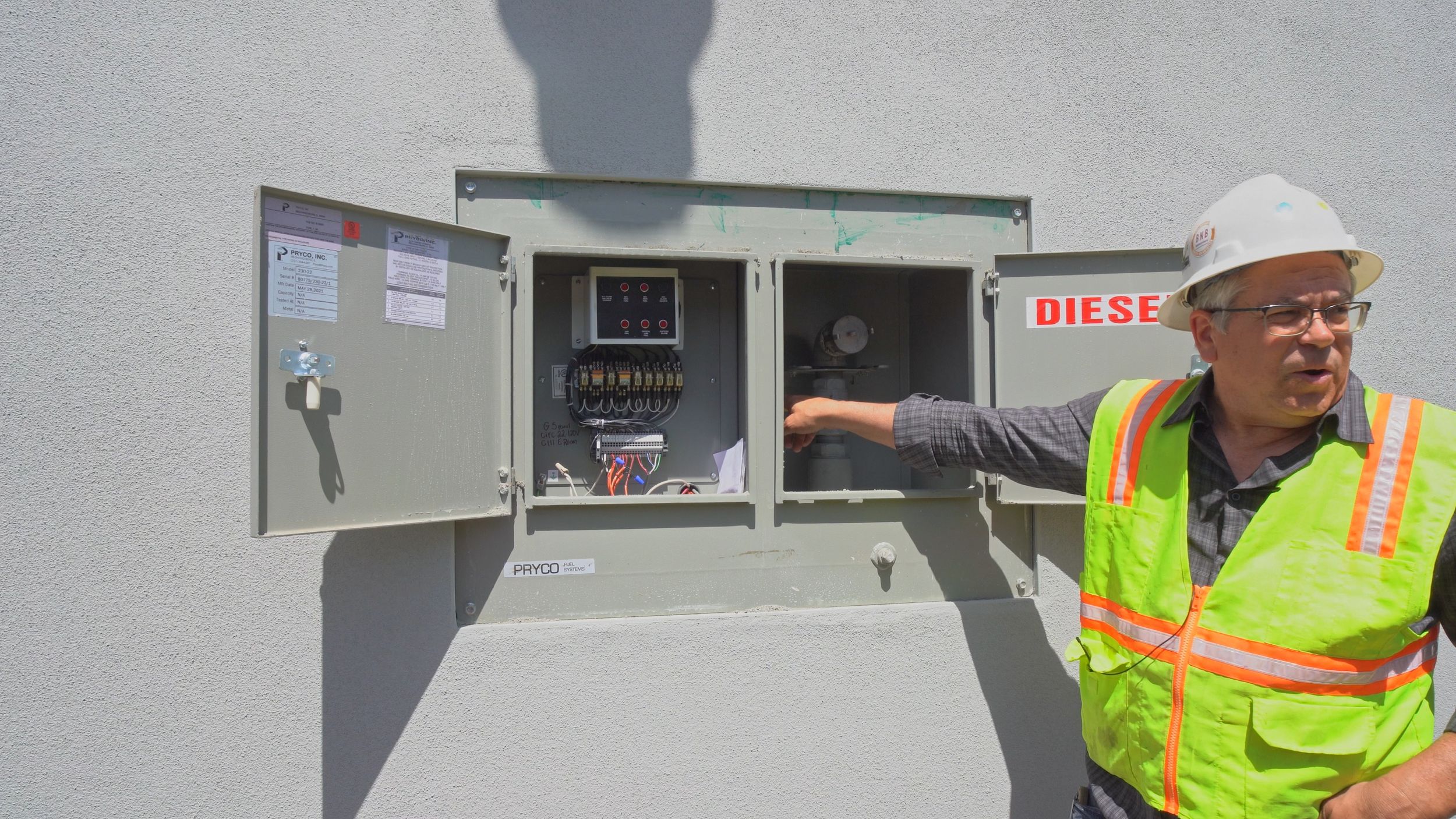 Industrial worker in a hard hat and safety vest pointing to the interior components of a wall-mounted commercial diesel control box.
