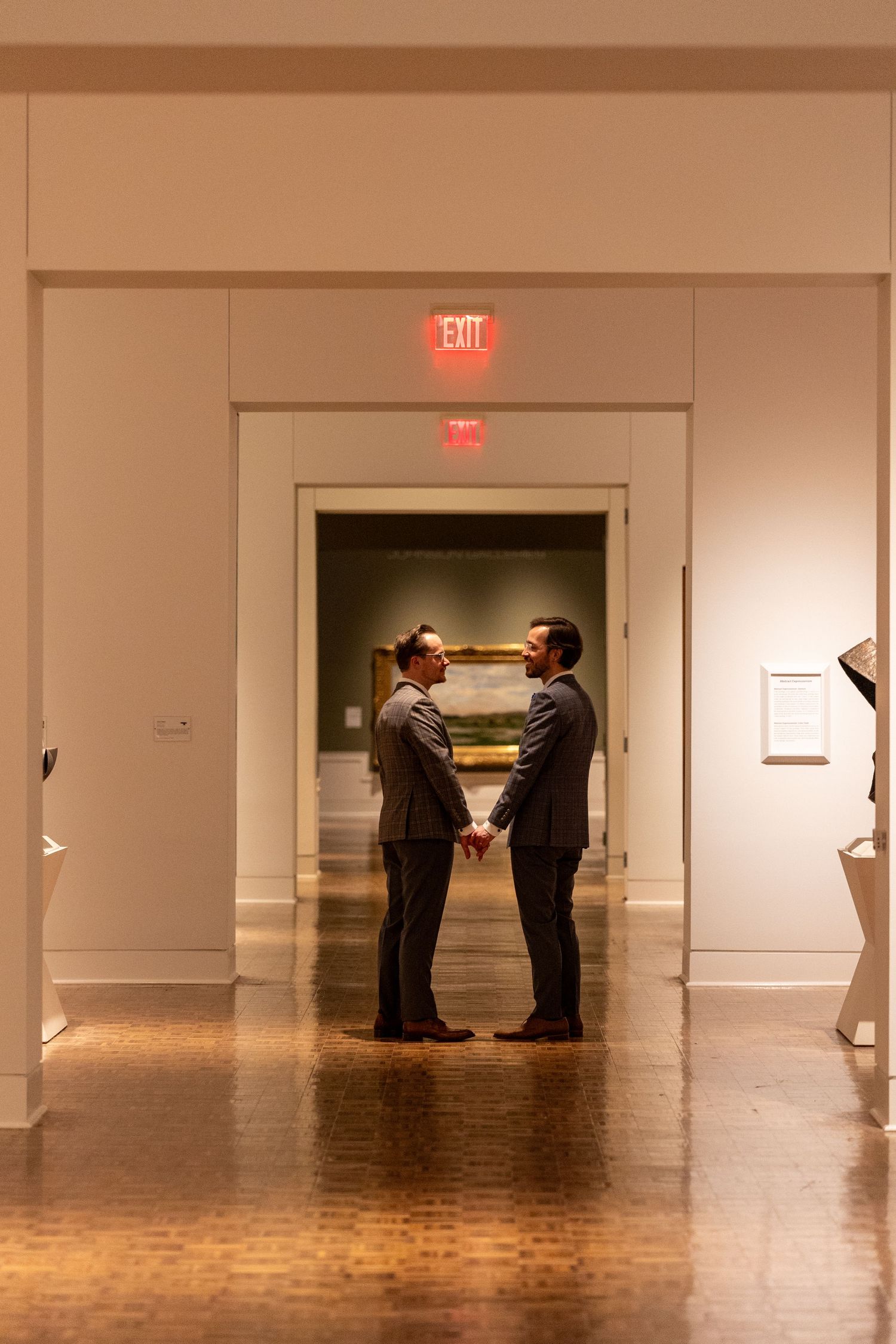 Two grooms in suits holding hands in a warmly lit museum hallway during their wedding ceremony.