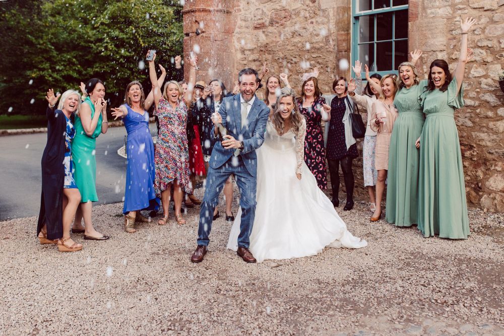 Wedding party and guests gather outside a historic stone building for group photos.