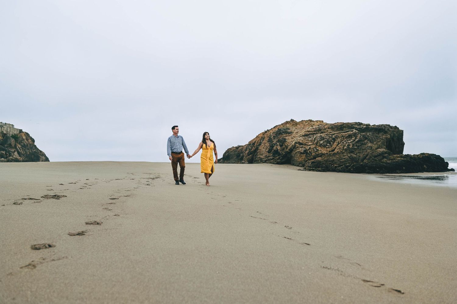 A couple walks along a scenic beach with rocky outcrops in the background.