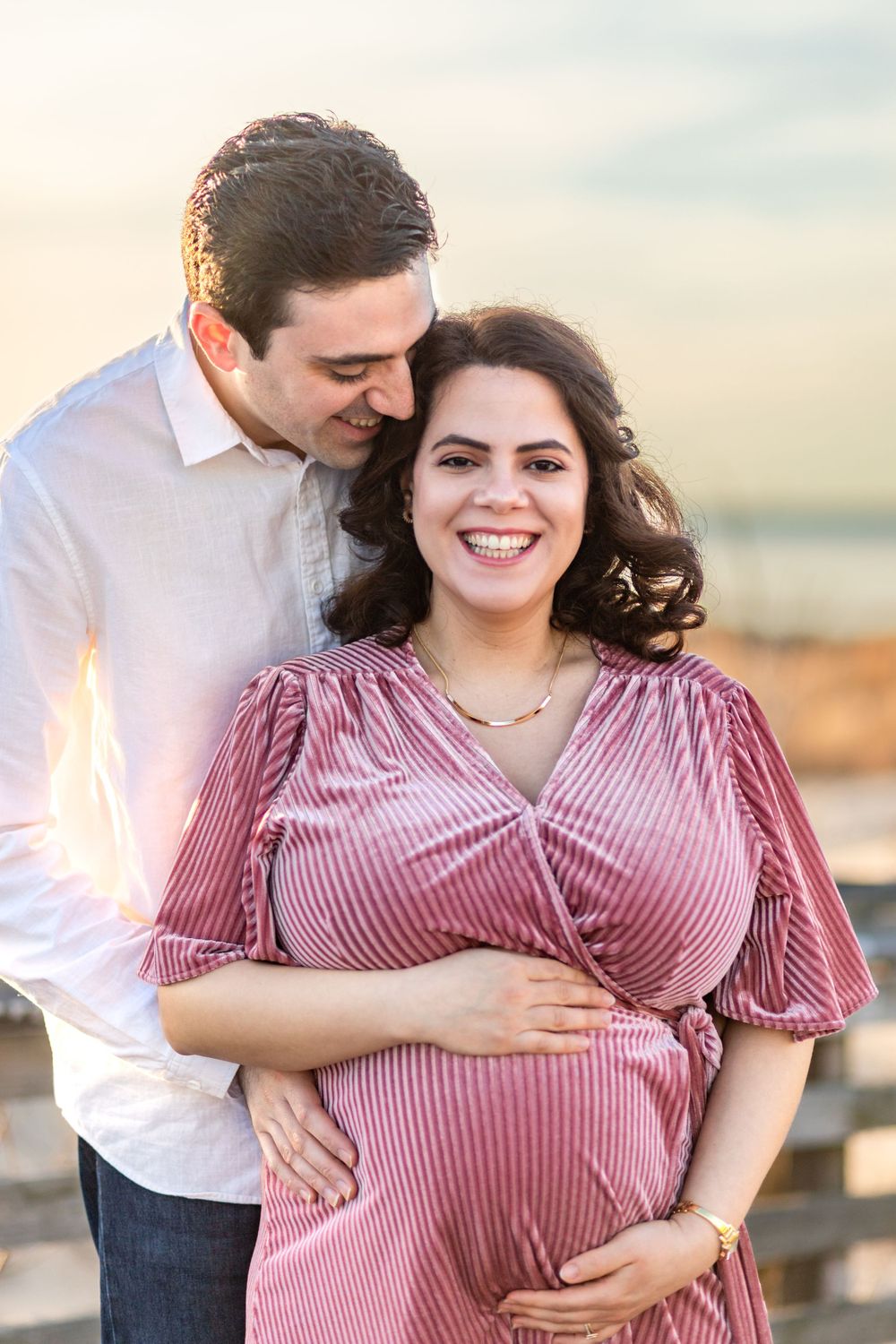 A couple shares a tender moment at sunset with the woman wearing a pink velvet maternity dress.