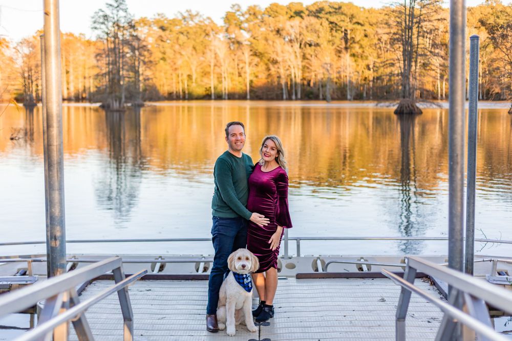A couple poses with their dog on a wooden dock overlooking a golden sunset lake.