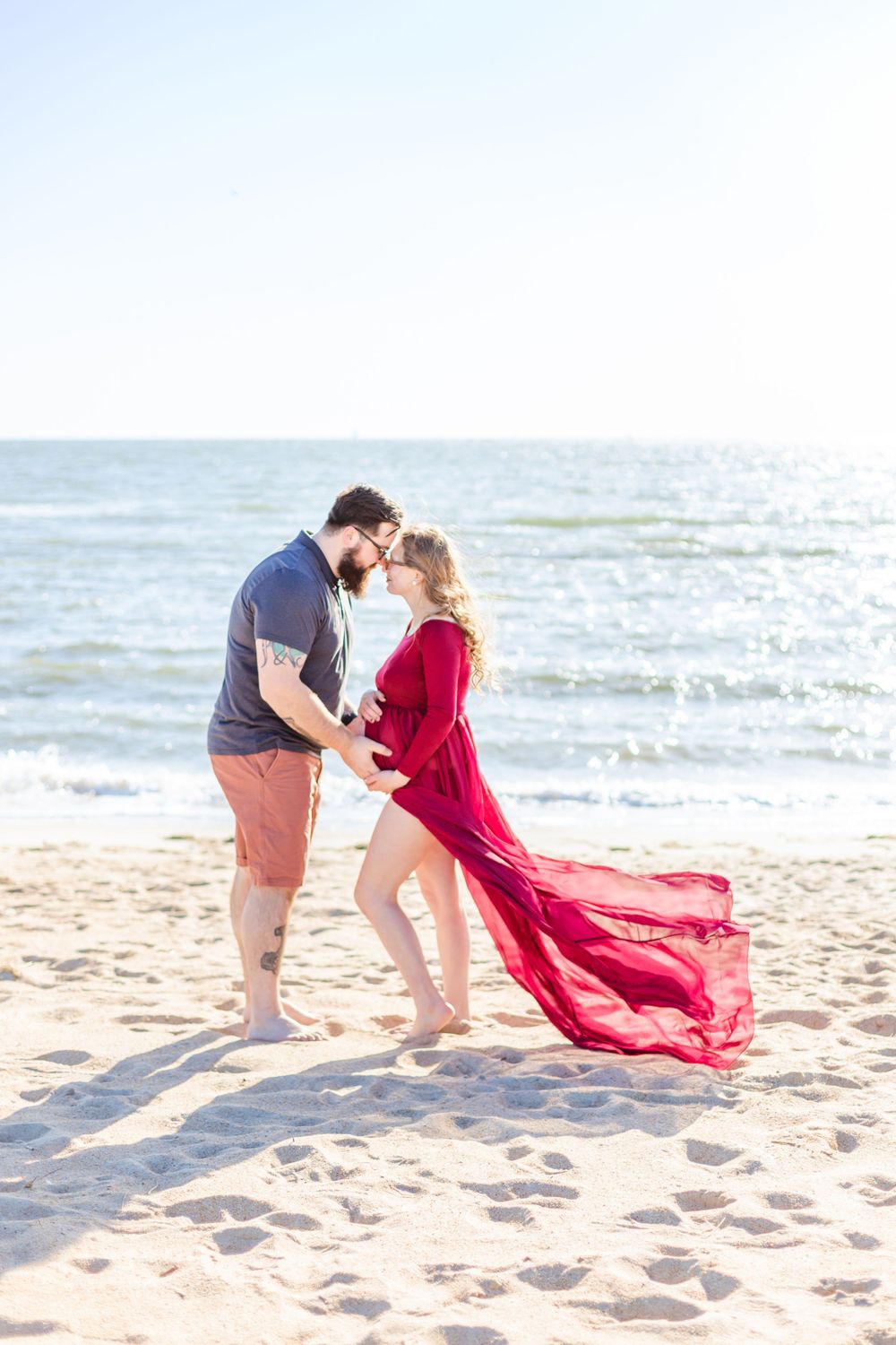 A couple embraces on a beach with a flowing red dress catching the wind.