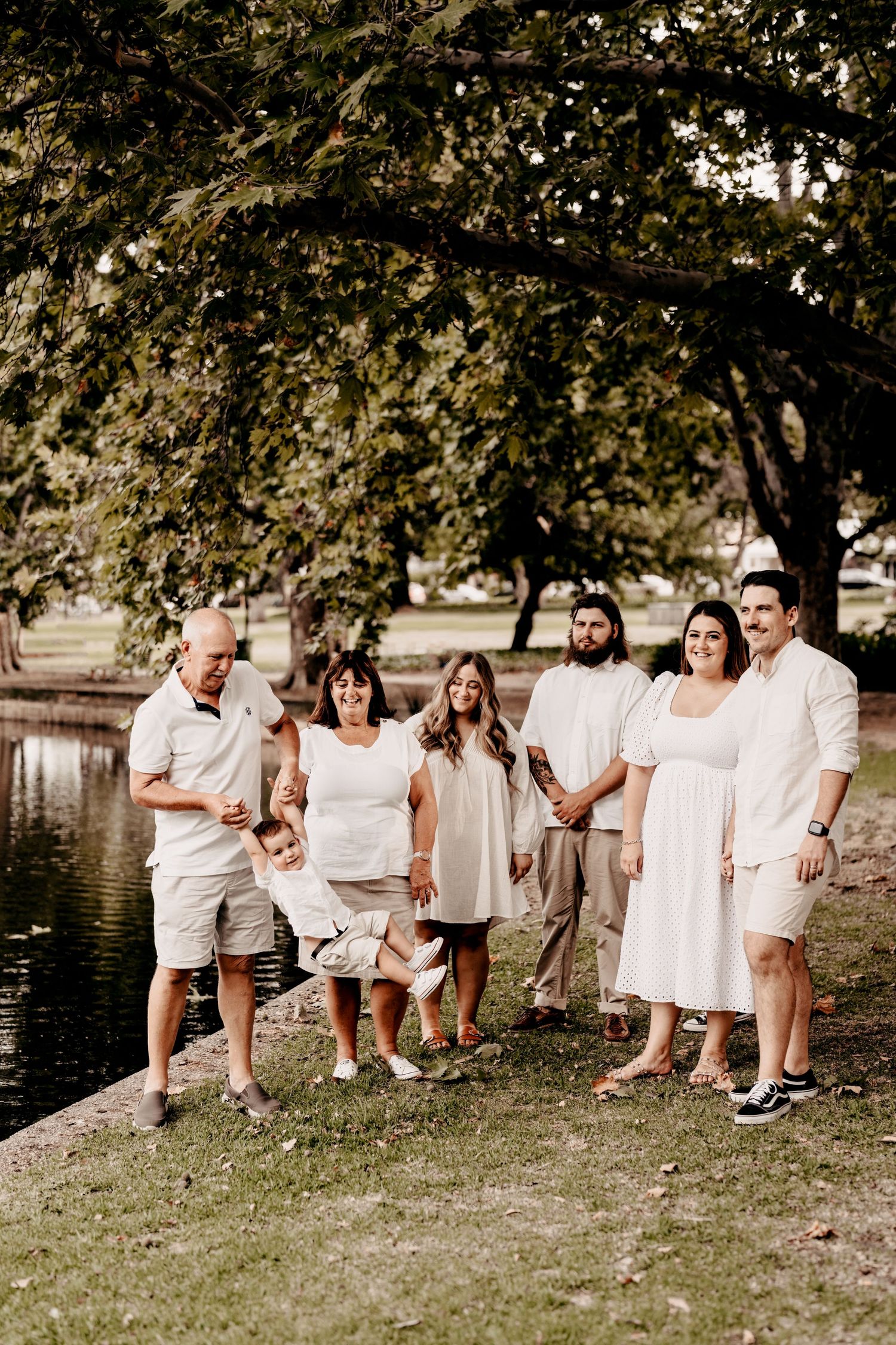 Extended family in white clothing sitting together by a lake under tree branches.