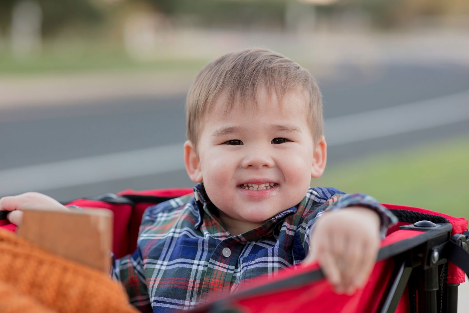 A child wearing a plaid shirt sits in a red wagon outdoors with a cheerful expression.