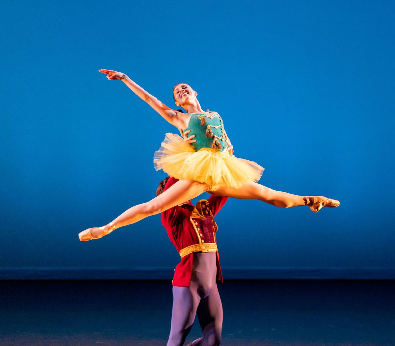 Ballet dancers perform dramatic lift with yellow tutu against blue stage backdrop.