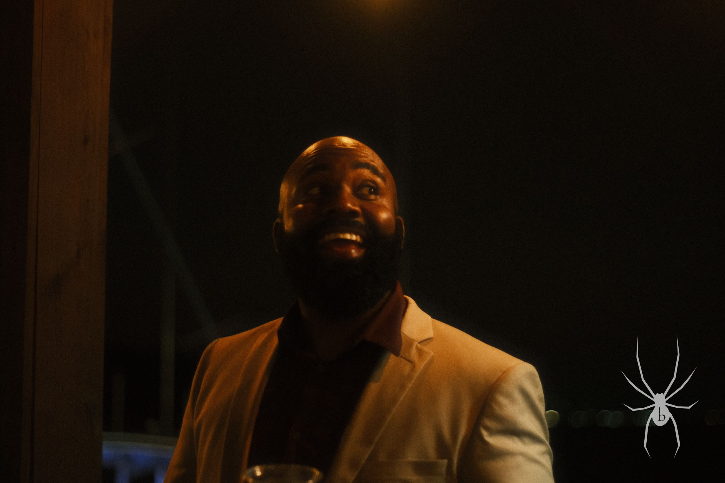 Low-lit portrait of a bearded person in formal jacket against dark background.
