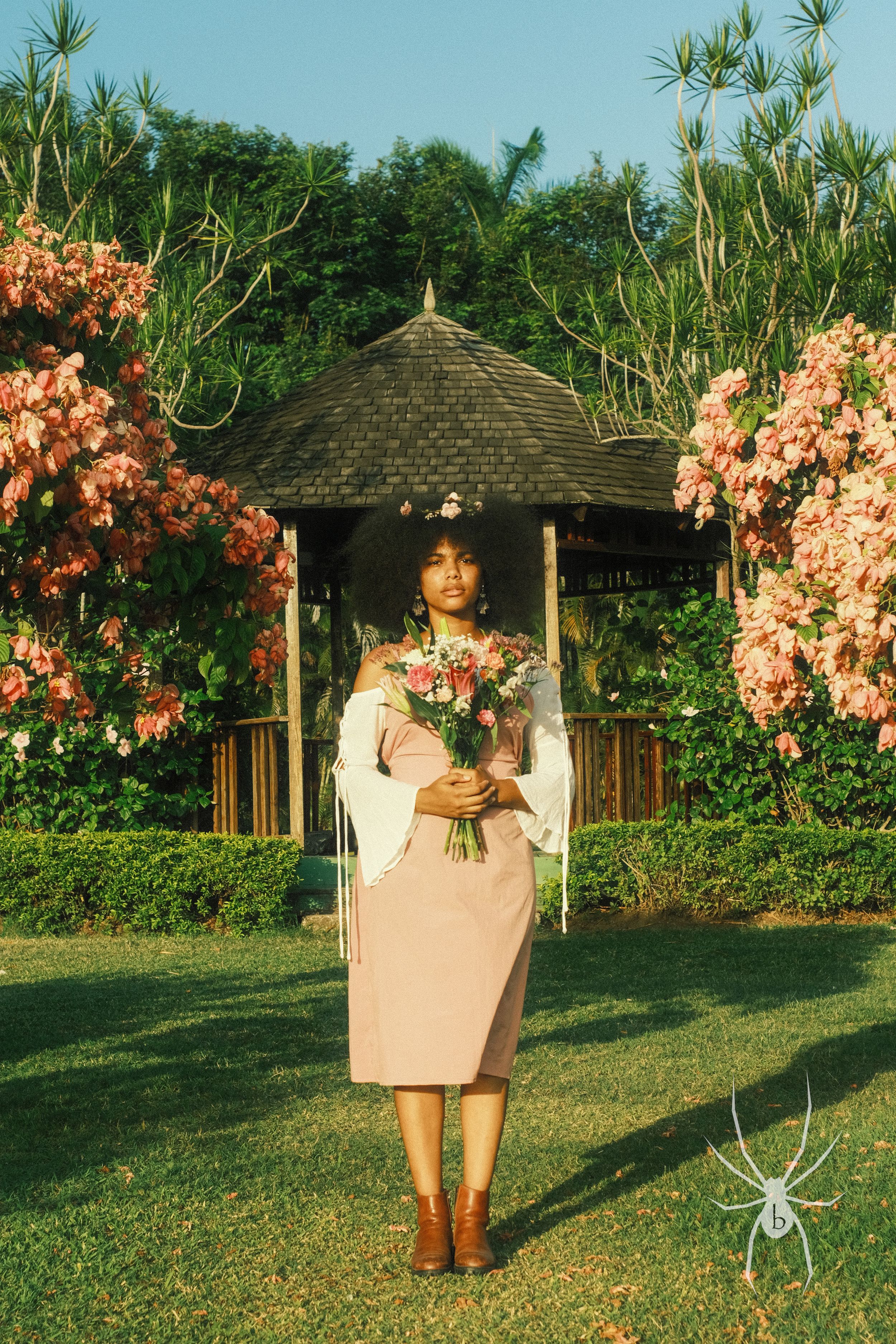 Person holding flowers in pink dress by gazebo with coral blooms and spider watermark.