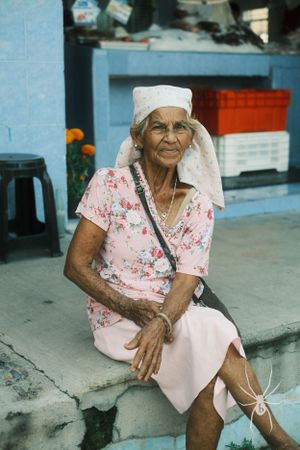Elderly woman in floral dress and white headscarf sitting on concrete step.