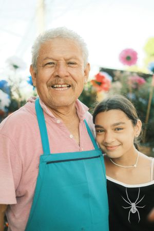Two people smiling together at outdoor event, one wearing turquoise apron and pink shirt.