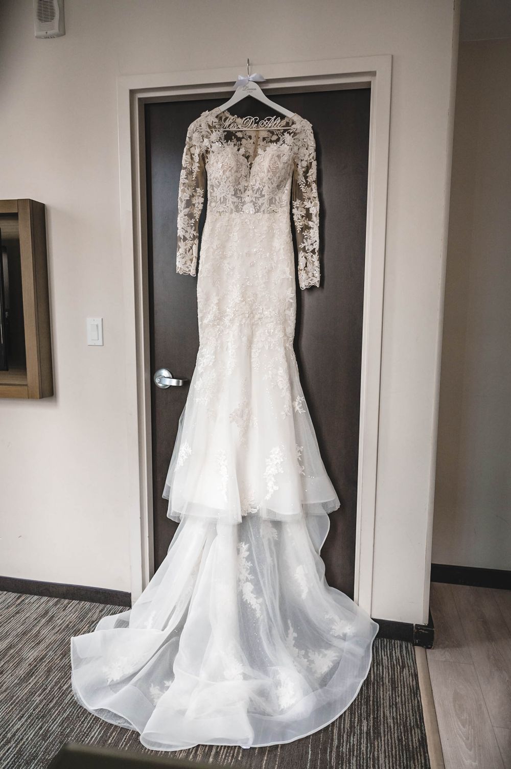 Elegant white lace wedding dress displayed on a hanger against grey walls.