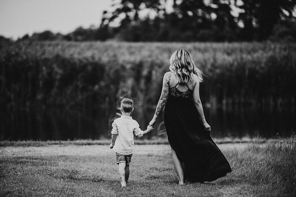 A person in a flowing black dress walking hand in hand with a small child in a field at sunset.