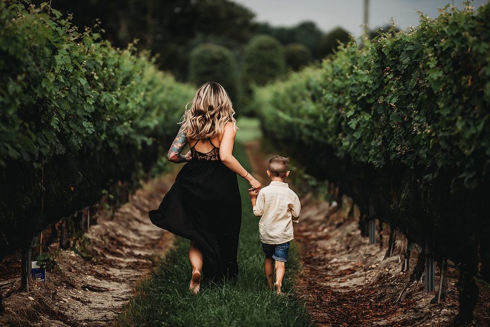 Running through vineyard rows in black dress with child on sunny day.