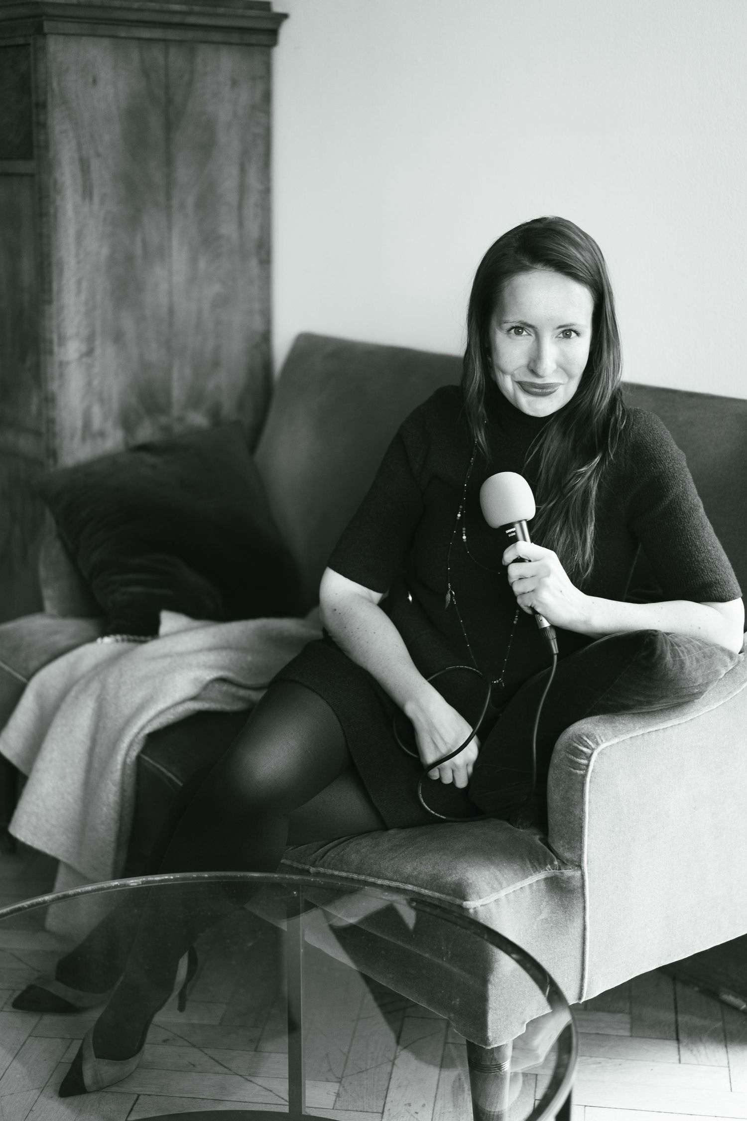 Woman in black outfit holding microphone while sitting casually on gray couch.