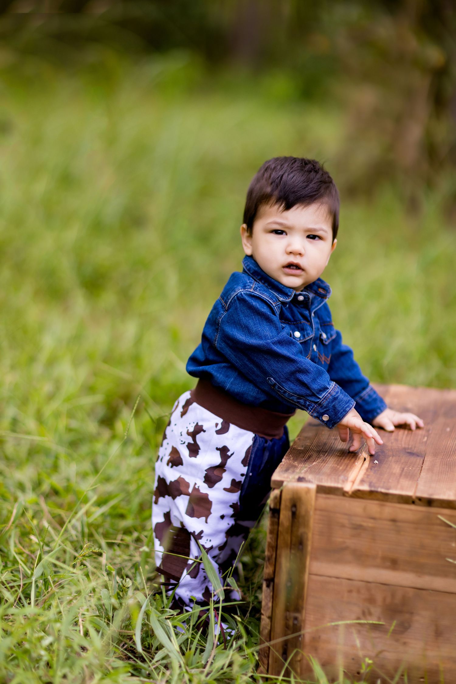 Small child in blue shirt and patterned pants stands by wooden crate in grassy setting.