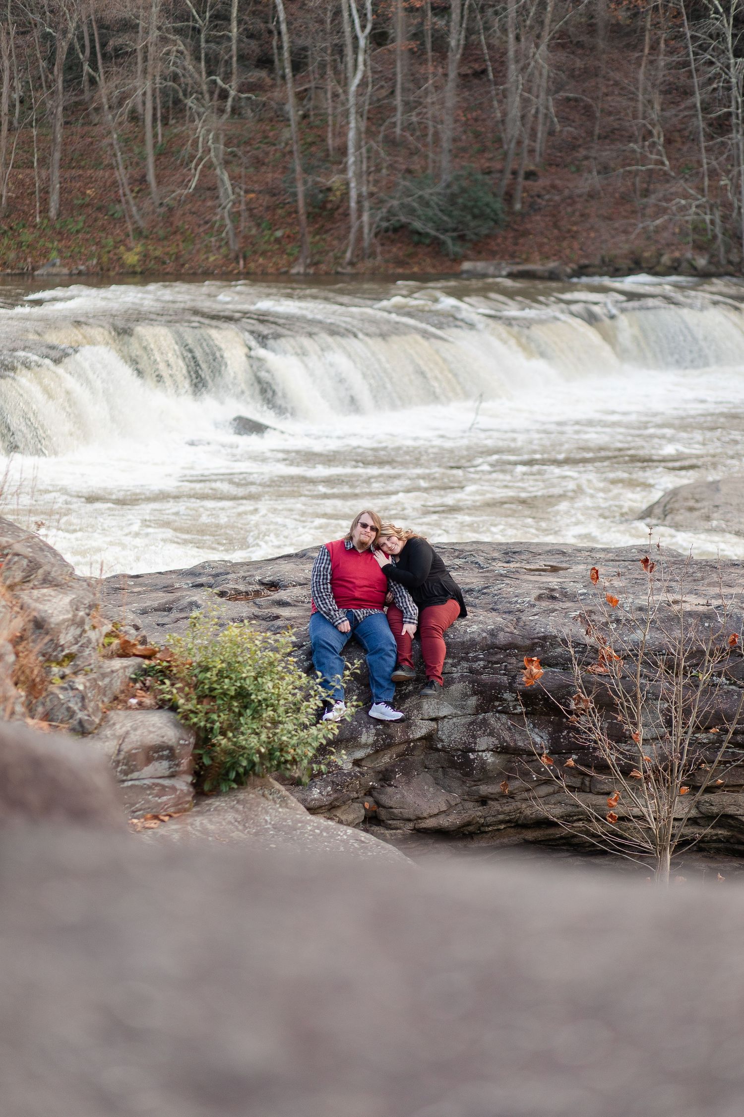A couple sits together on rocks overlooking a rushing waterfall in a wooded setting.