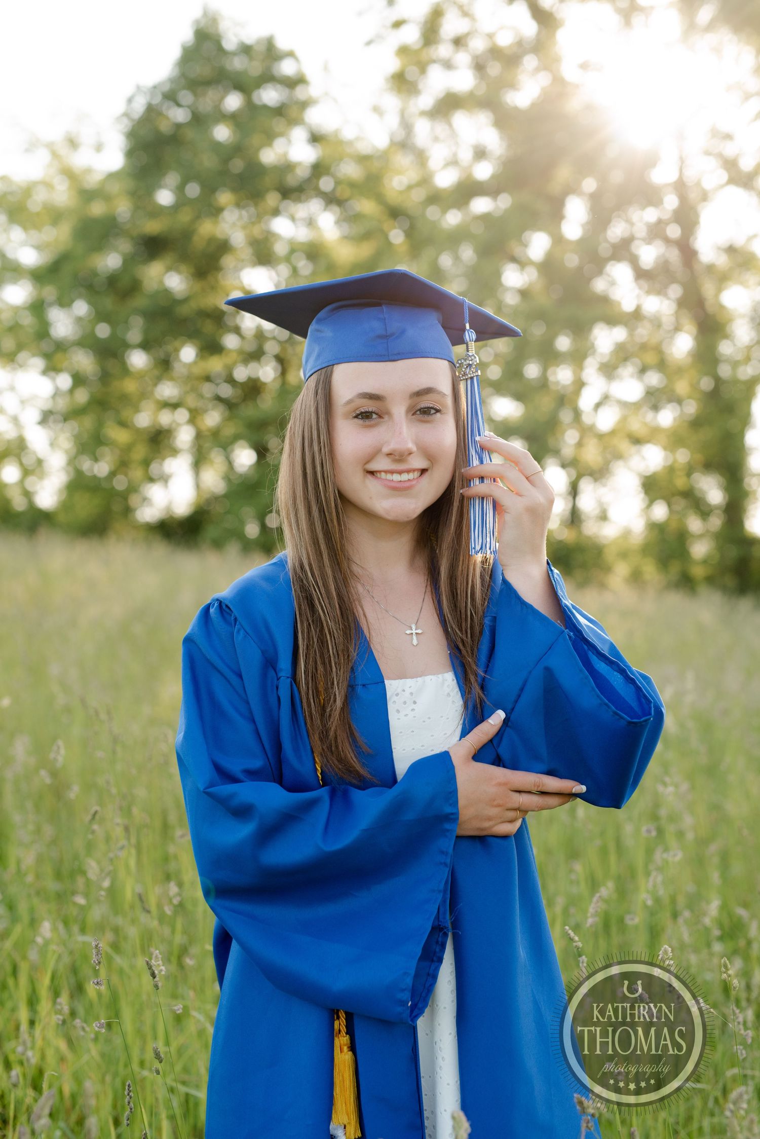 Graduate wearing bright blue gown and cap poses in sunny field with tassel.