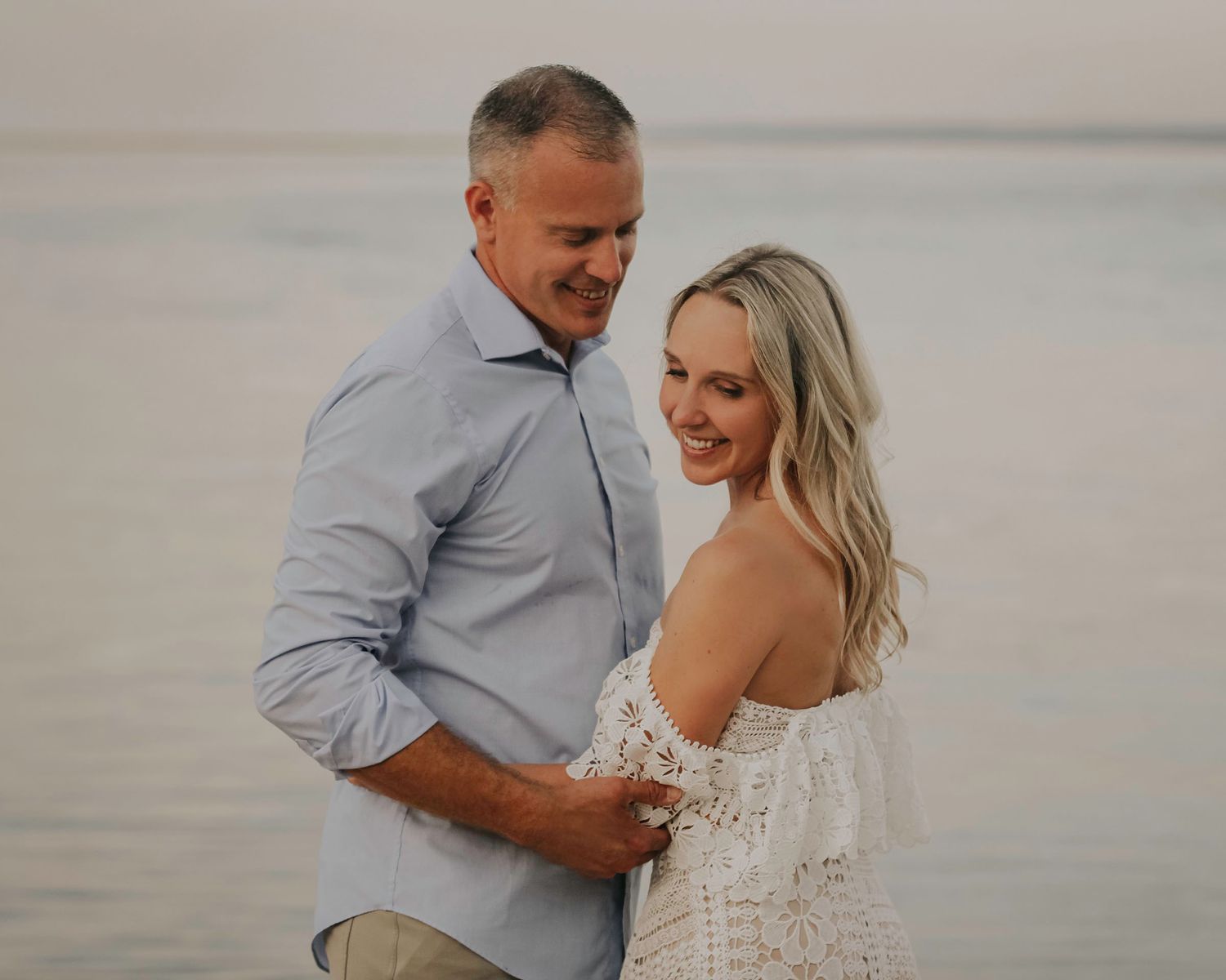 Romantic beachside photo series with couple embracing at sunset in soft muted tones.