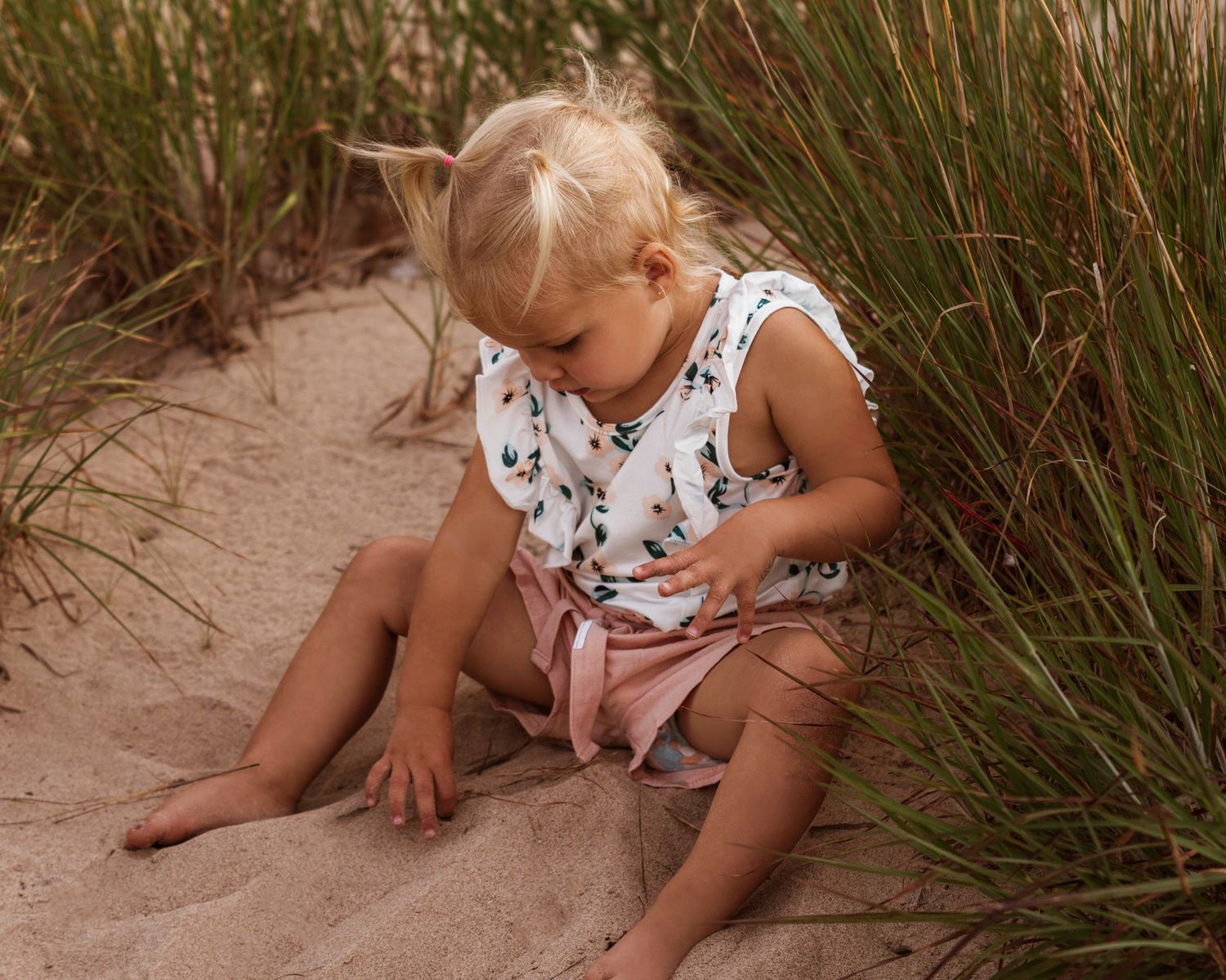 A child in a white floral dress sits on sandy ground surrounded by beach grass on a sunny day.