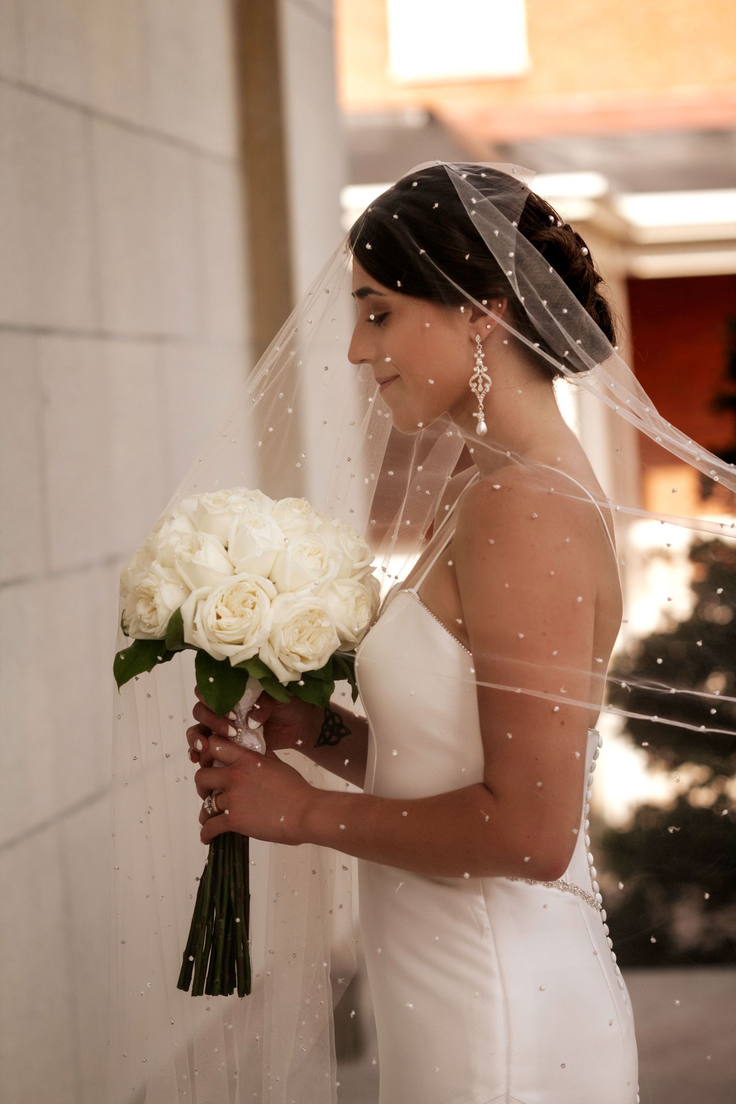 A bride in a sleek white gown holds a bouquet of white roses while wearing a pearl-dotted veil.
