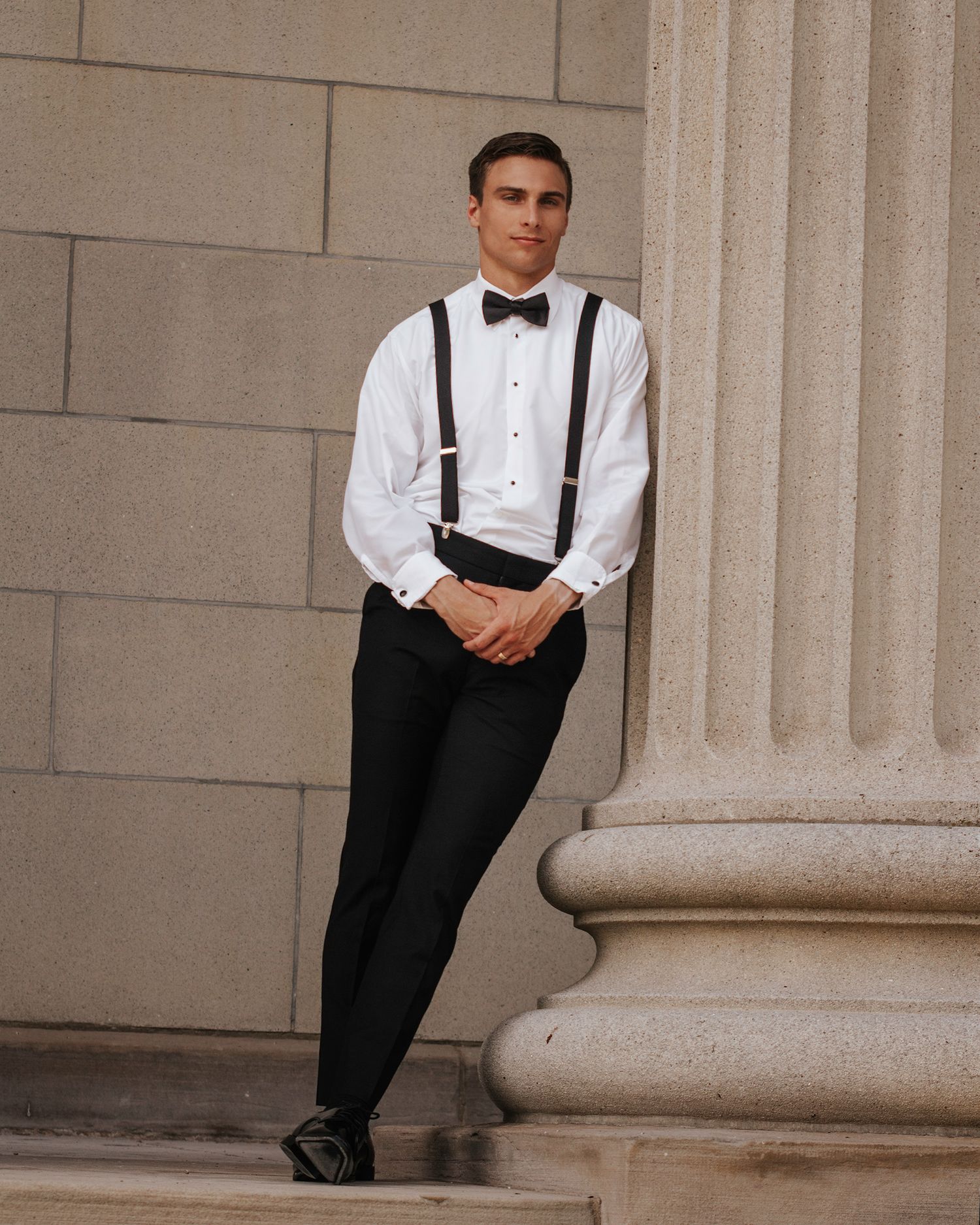 A formal portrait of someone in classic black and white attire with suspenders against a stone column.