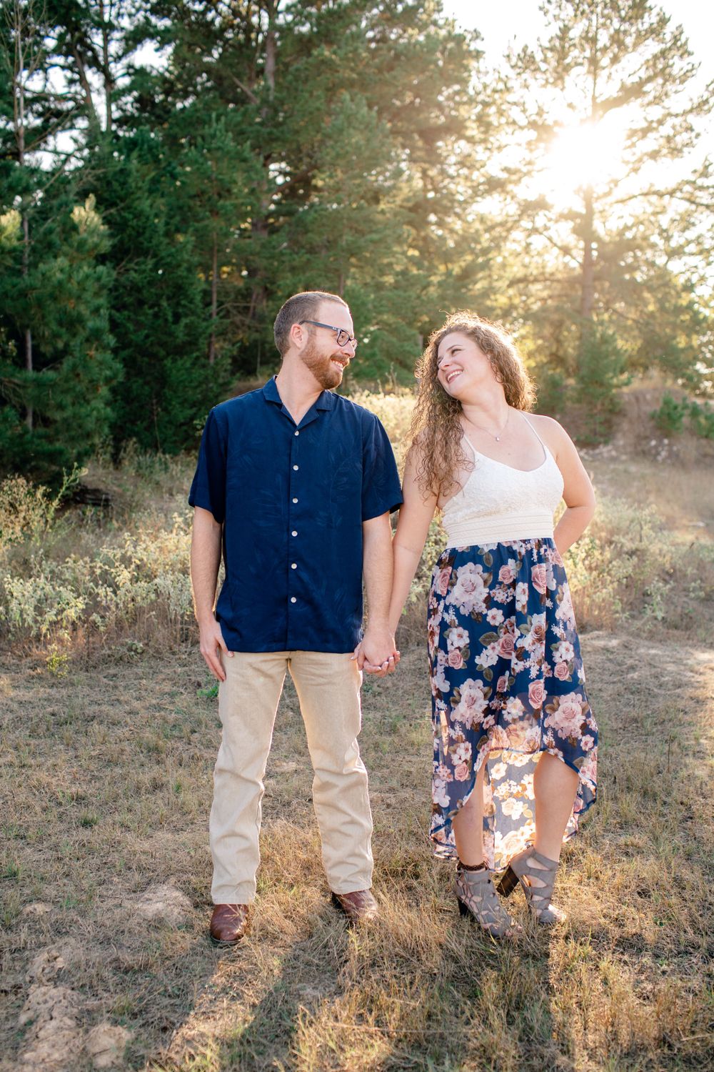A couple laughing together in a sunny forest setting wearing casual summer outfits.