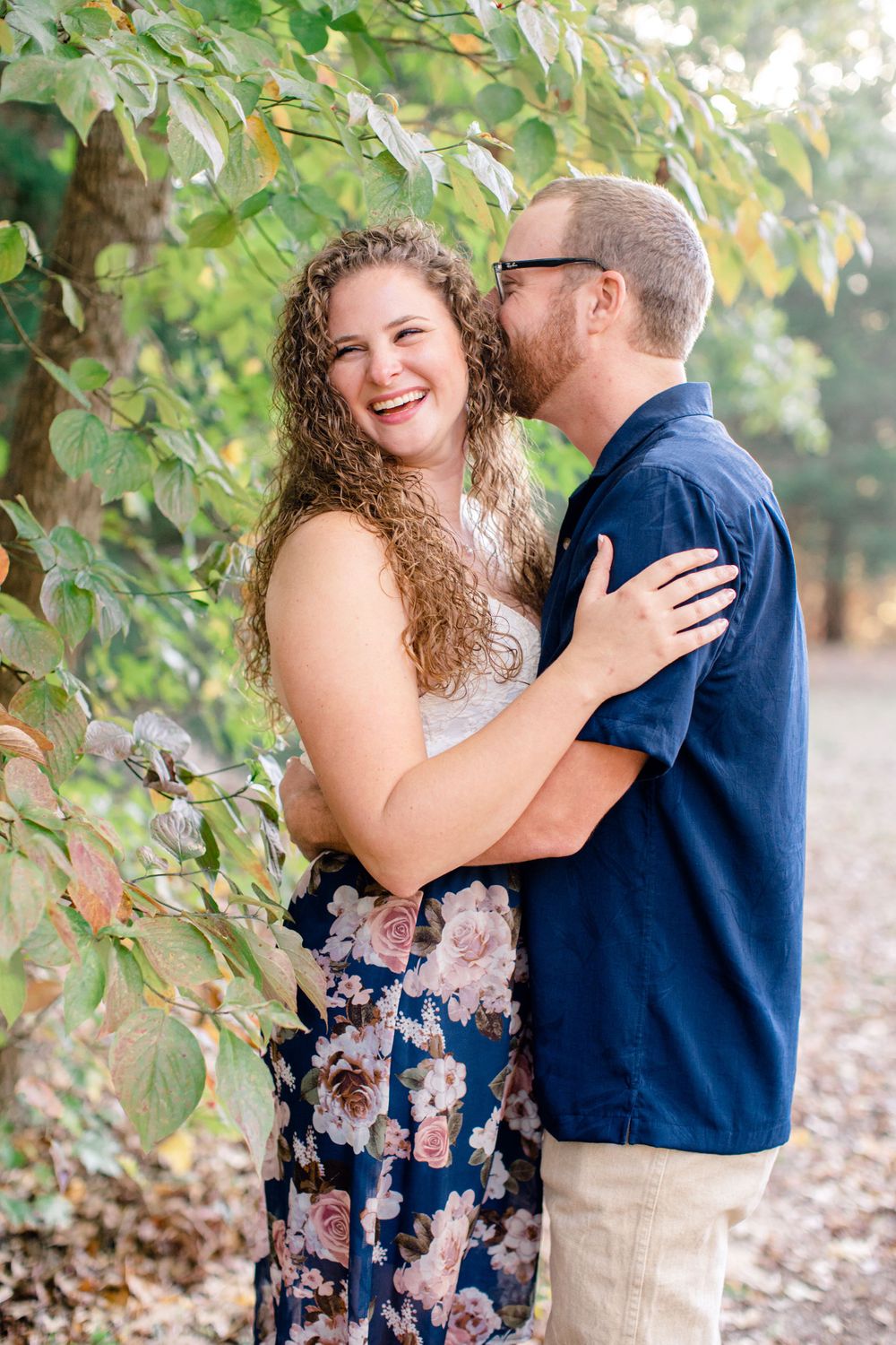 Romantic outdoor portrait of a couple embracing under green leaves in golden sunlight.