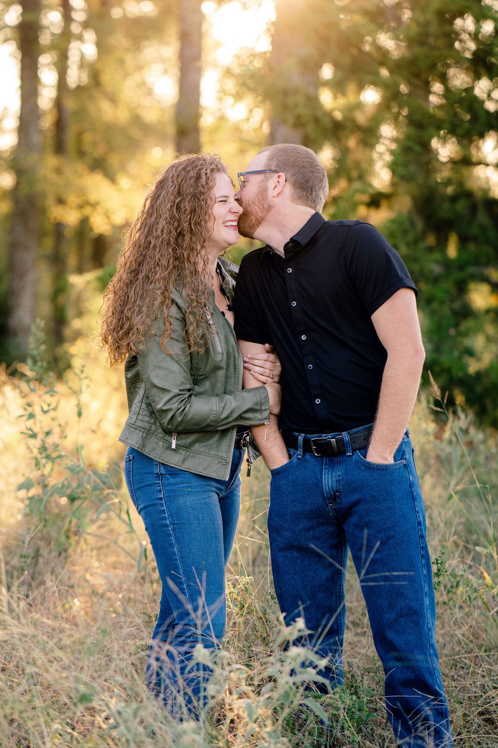 Couple sharing an intimate moment while standing in tall grass with sunlit trees in background.