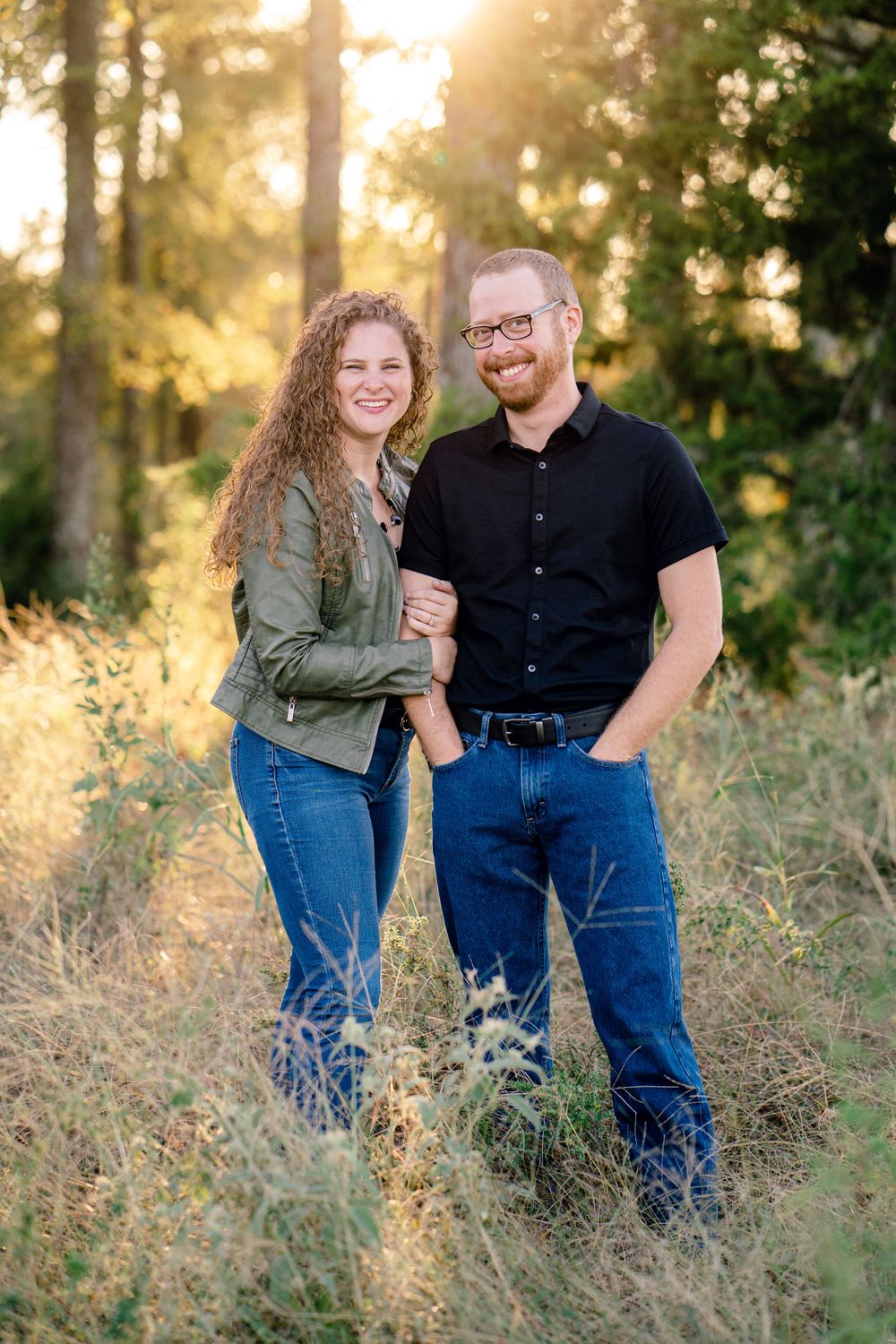 Happy couple poses together in casual attire against a sunlit forest backdrop.