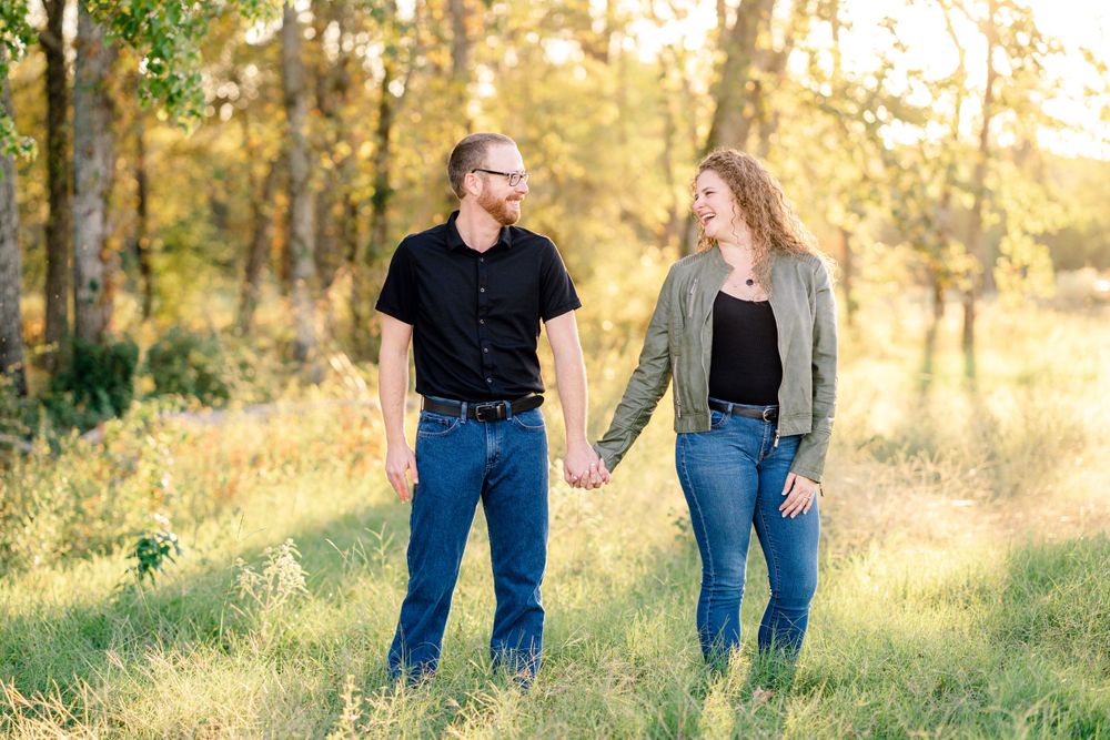 Couple holding hands while walking through a sunlit meadow with trees in background.