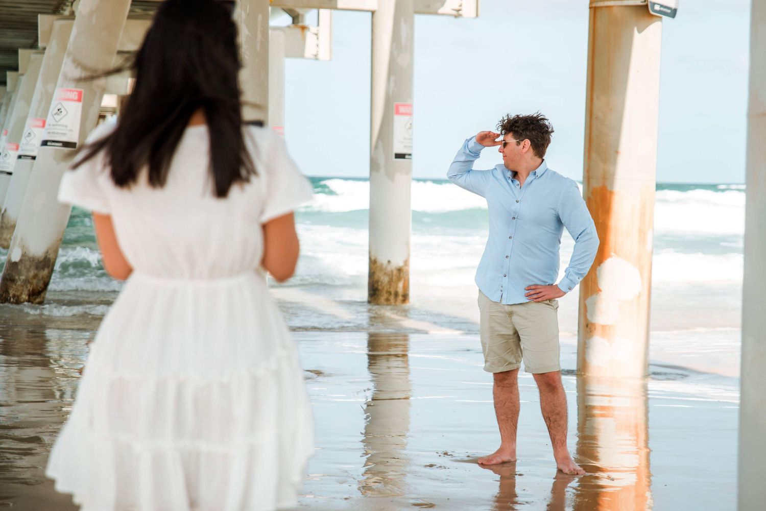 A romantic scene under a pier with waves and a couple in summer outfits.