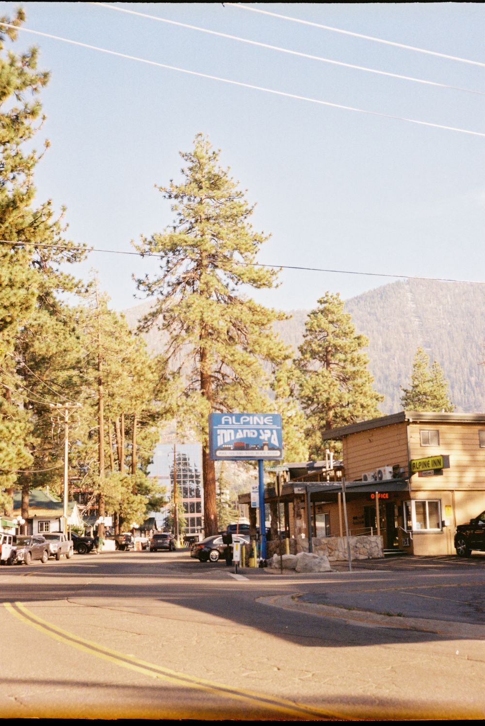 A small town street corner with tall pine trees and mountain views in the background.