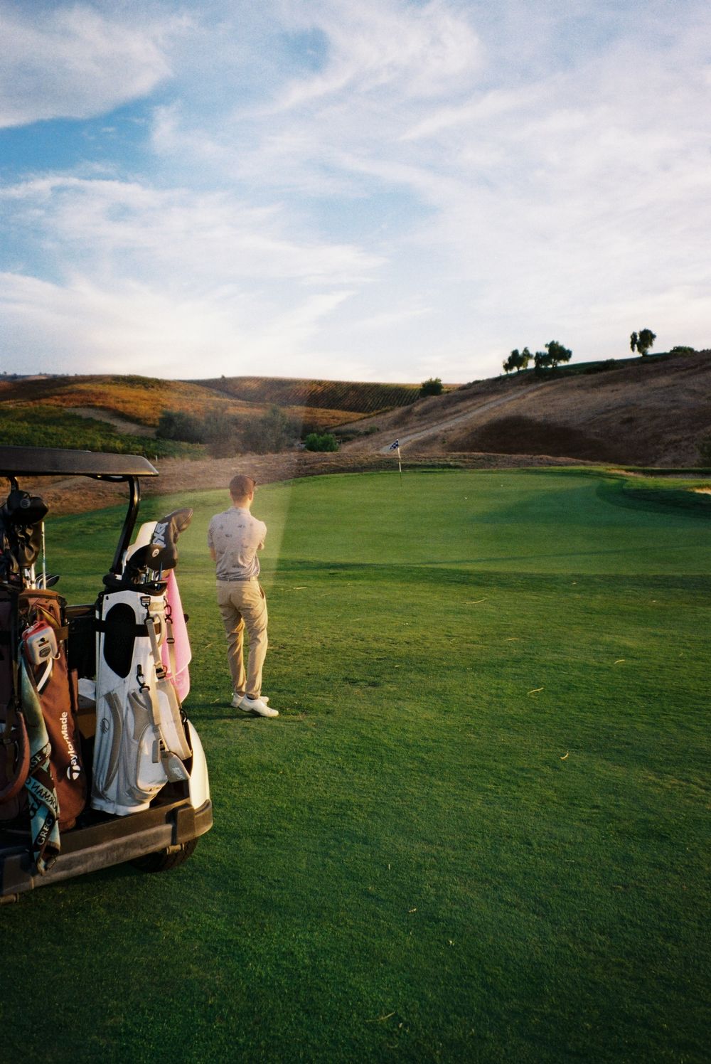Golf cart and players on lush green course with rolling hills in background.