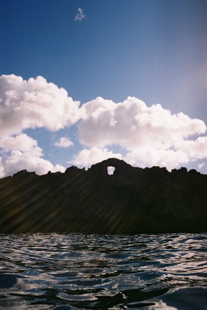Dark silhouette of a mountain with a hole against cloudy sky and rippling water.