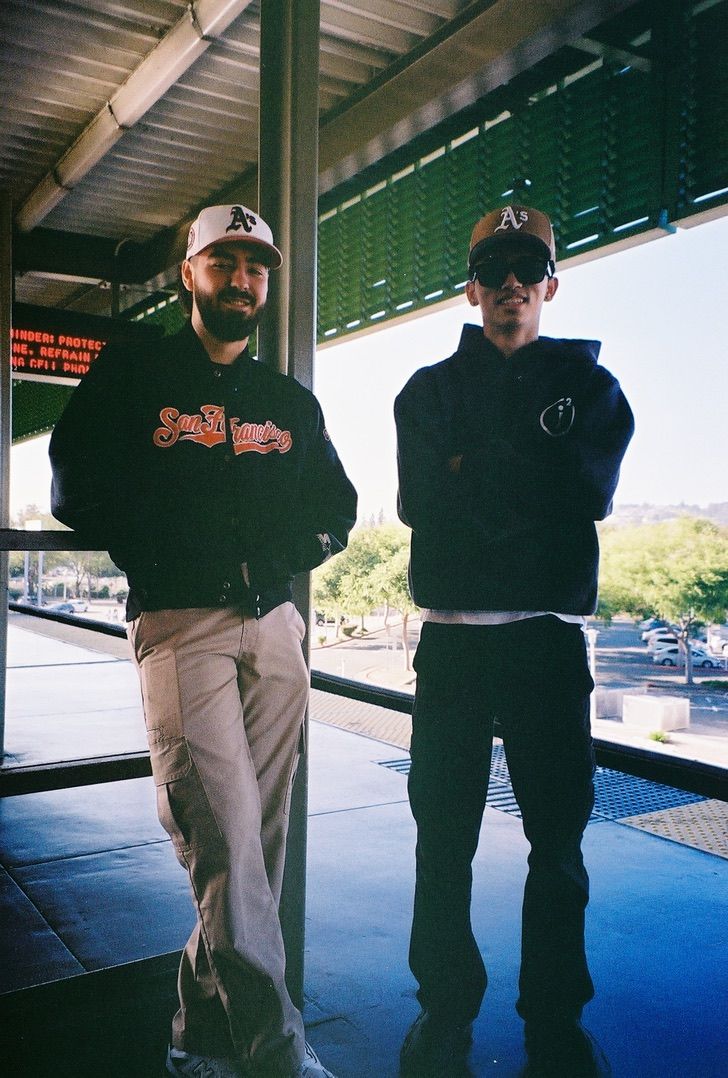Two people wearing baseball caps stand in casual attire at what appears to be a sports venue.