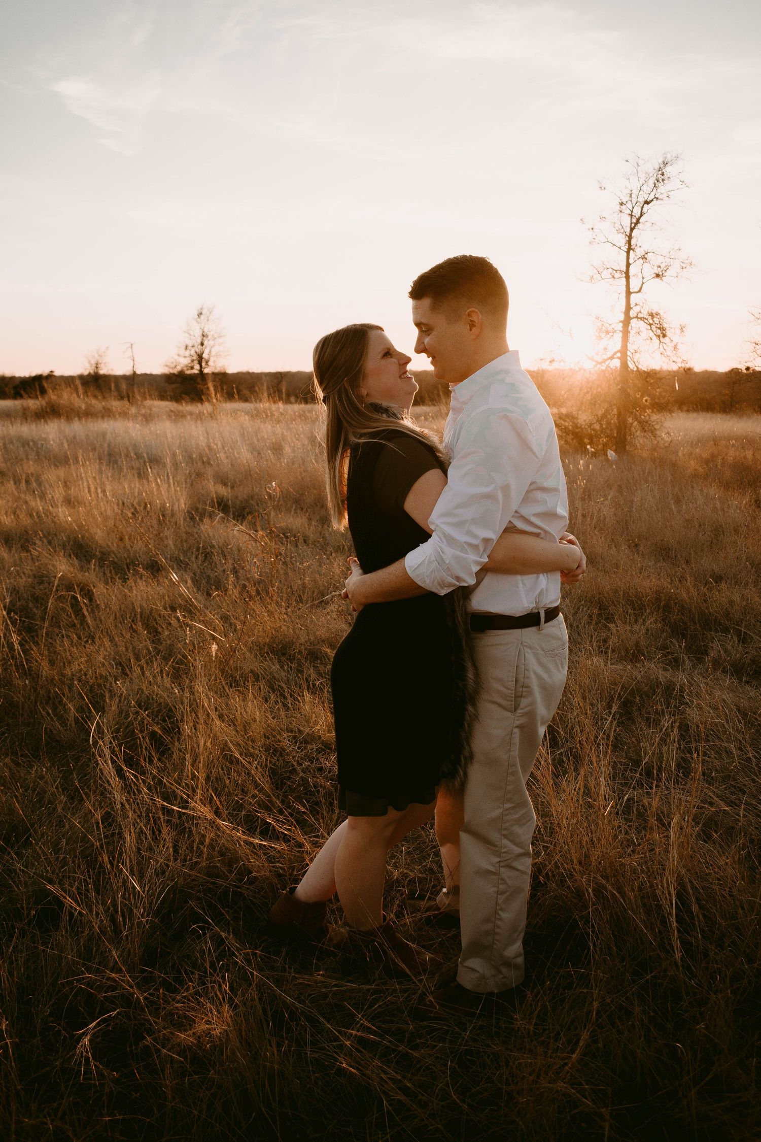 A romantic couple embraces in a golden-lit field during sunset with dry grass surrounding them.