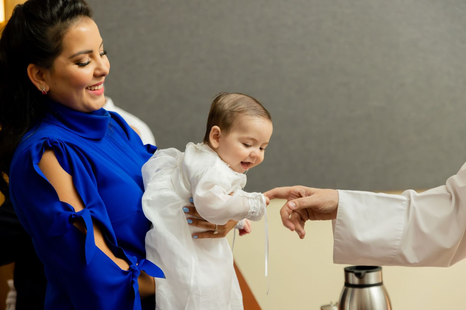 Woman in blue dress holding baby in white christening gown during baptism ceremony.
