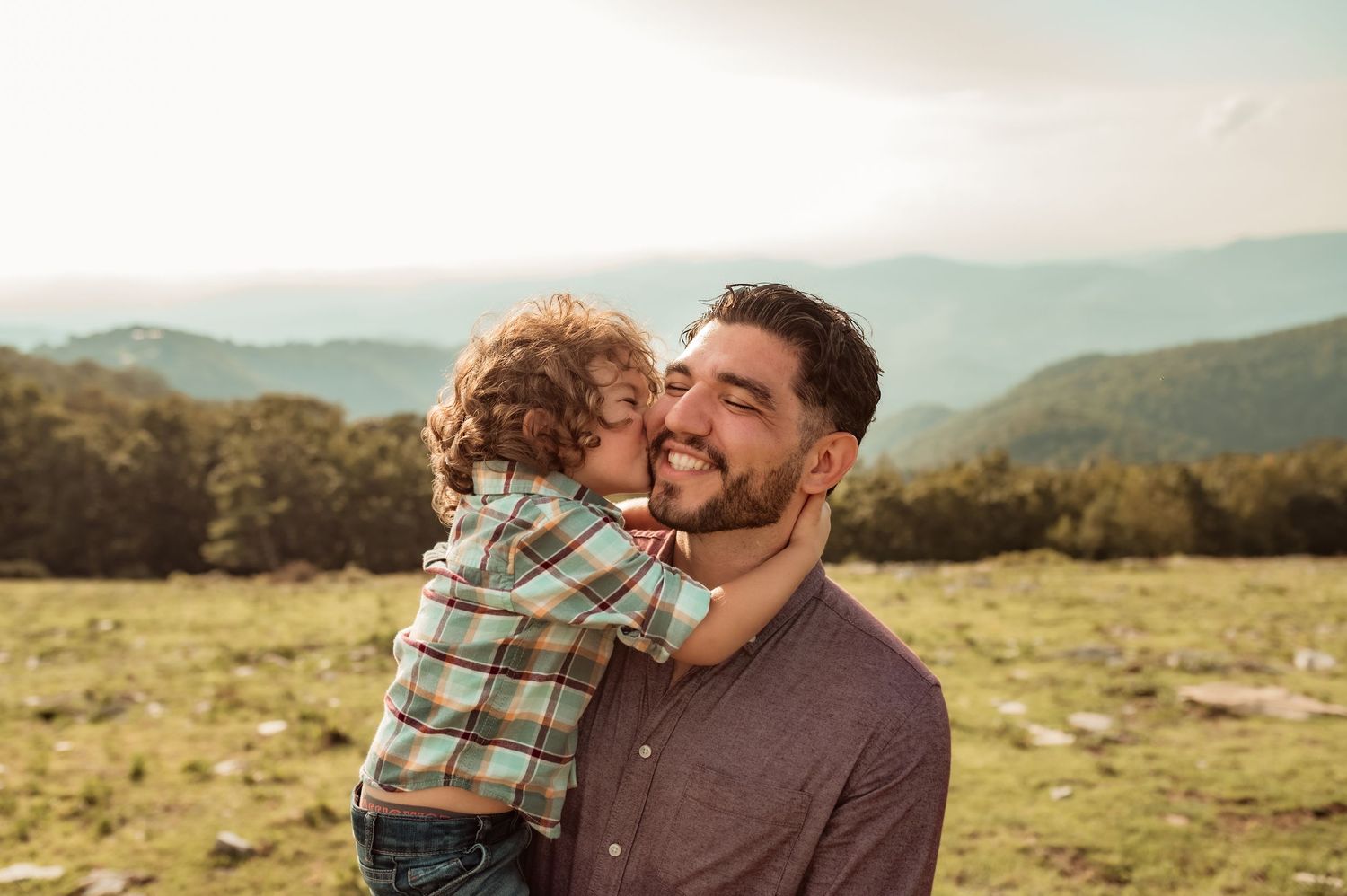 Dad smiles with eyes closed while son kisses him on cheek with arms wrapped around his neck lovingly.